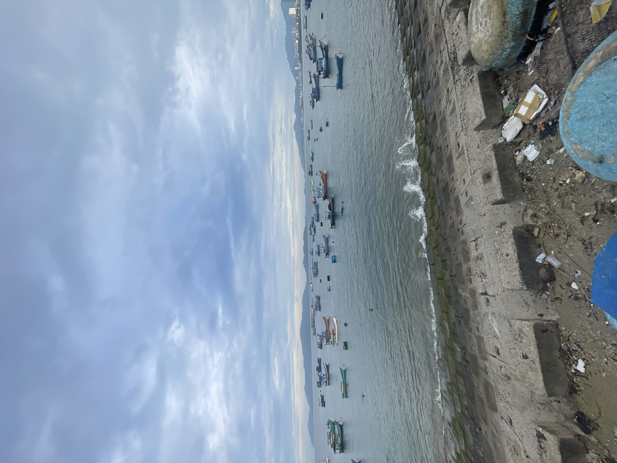 The image captures an overcast coastal scene, likely a fishing harbor or bay, under a cloudy, light grey sky that fills the upper portion of the frame. In the middle ground, a wide expanse of calm, grey-blue water is dotted with numerous small fishing boats. These boats vary in color and design, with many exhibiting characteristics of traditional Vietnamese vessels, and appear to be either moored or at anchor.
On the distant horizon, beyond the water and boats, several layers of mountains rise, partially obscured by a light haze. The faint outline of a city skyline with taller buildings can also be seen in the distance against the mountains.
In the immediate foreground, a concrete seawall or embankment runs along the edge of the water. The wall is constructed from large, interlocking concrete blocks, some of which are stained green with algae where small waves gently lap against them. The top of the embankment is cluttered with various debris and trash, including discarded plastic items, styrofoam, and general refuse. Two large, blue, bowl-shaped objects, possibly traditional Vietnamese coracle boats or large containers, are prominently positioned on this cluttered ground in the foreground.
The time of day appears to be daytime, likely late morning or afternoon, given the soft, diffused light from the cloudy sky. No people are visible, and there is no discernible text in the image. The scene portrays a tranquil yet somewhat neglected coastal area, highlighting local fishing activity alongside visible environmental debris.