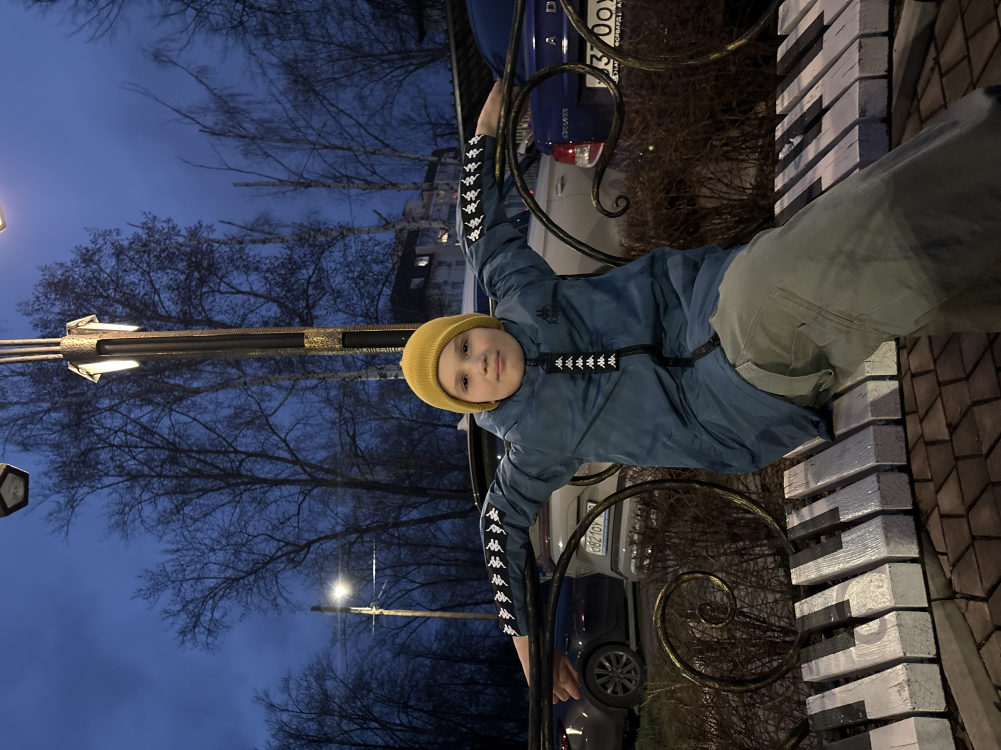 This image, taken in Chaikovsky, Russia, depicts a young boy, approximately 5-8 years old, posing outdoors during dusk or early evening. The sky is a deep blue, and the bare branches of trees suggest a cold season, likely late autumn or winter. Artificial lights, including streetlights, are visible, illuminating the scene.
The boy is dressed in a yellow knitted beanie, a blue jacket featuring multiple "Kappa" logos along its sleeves and on the chest, and light-colored pants. He is holding onto a decorative, dark metal fence or railing with both hands, looking directly at the camera with a calm, slightly smiling expression.
In the background, two cars are parked. A blue Lada Granta is visible, displaying "GRANTA" and "ФОРВАРДА АТ" (likely part of a dealership name) on its rear, along with a license plate showing "X300AD". Another lighter-colored car has a partially visible license plate, with the sequence "0821OY" identifiable through optical character recognition. To the boy's right, there is a low wall or structure constructed from painted wooden blocks, distinctively designed to resemble piano keys, with alternating black and white segments. The ground beneath is paved with brown pavers. The setting suggests an urban or suburban public space.