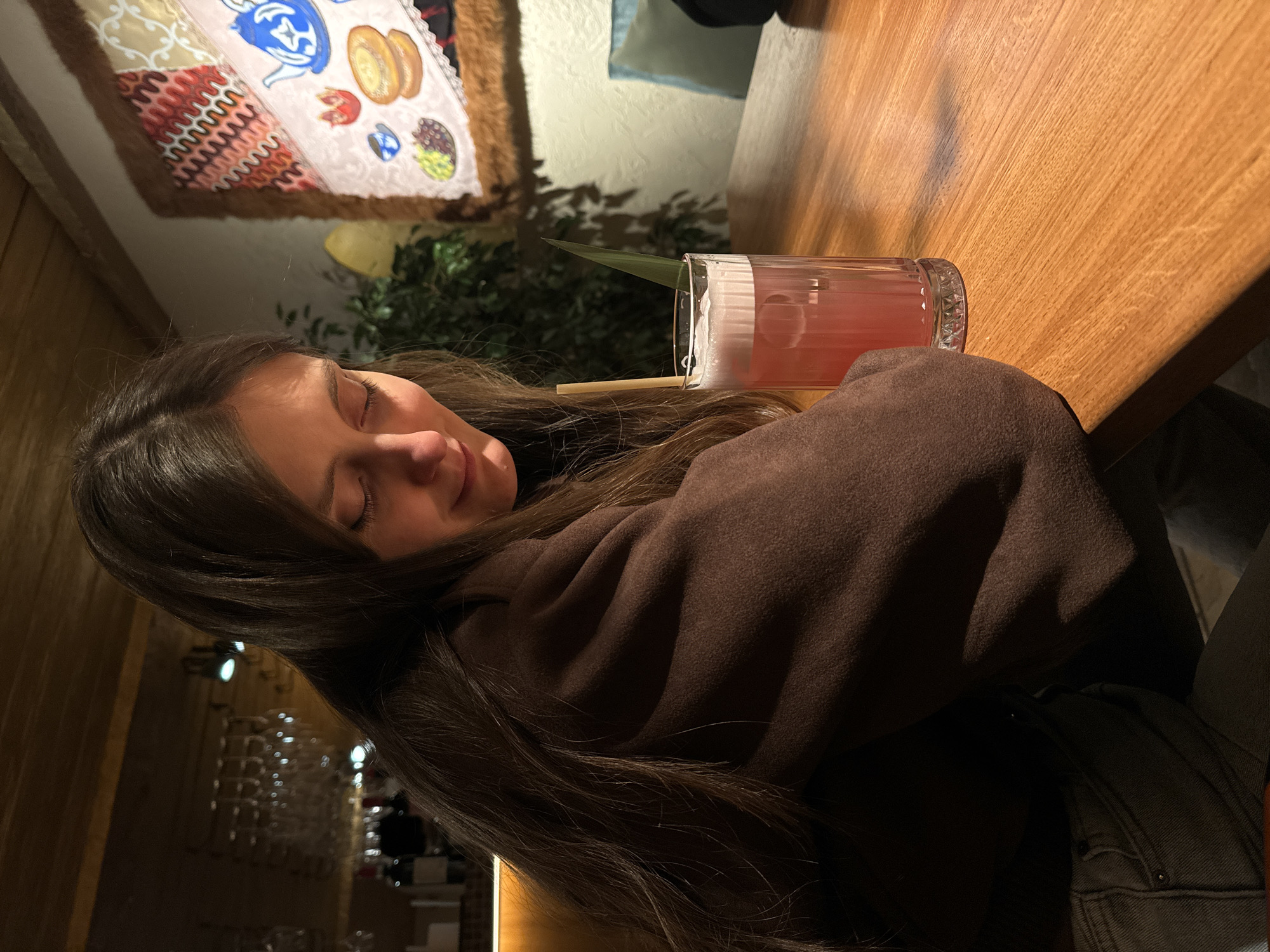A woman with long brown hair sleeps on a table next to a pink drink in a tall glass with a straw and a green leaf sticking out. The table is wooden, and the background includes a decorative wall hanging, plants, and a blurry view of a possibly upscale restaurant or bar. The location appears to be Moscow, Russia, given potential signage visible in the background, but this cannot be confirmed without additional imagery.