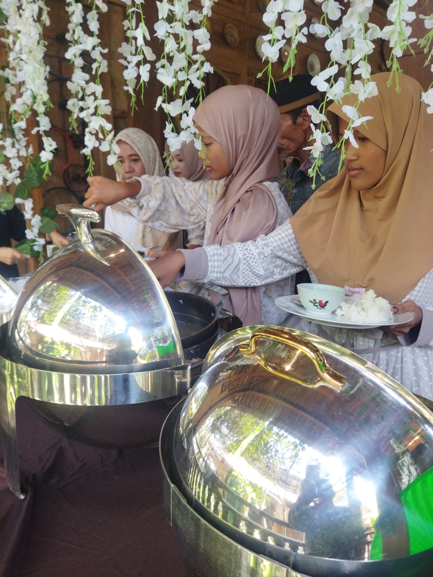 The image depicts a scene at a buffet, likely a wedding or celebration, in Semarang, Indonesia. Three women wearing hijabs are visible, serving food from large, stainless steel chafing dishes. The woman in the foreground, on the right, is holding a plate with white rice and a small bowl decorated with a red chicken motif. The woman in the middle is reaching into a chafing dish, and the woman on the left is also attending to the food. Behind them, a man in a dark cap is partially visible. The buffet is decorated with hanging white artificial flowers. The background suggests an outdoor or semi-outdoor setting with wooden walls and a thatched roof structure. The overall atmosphere is one of a communal meal service.