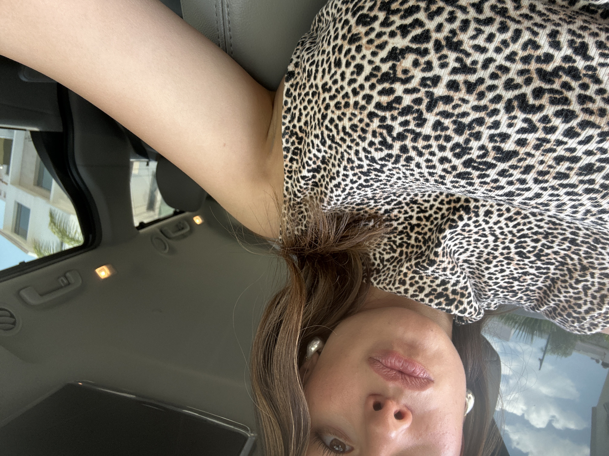 A woman is in a vehicle, wearing a leopard print top and silver earrings. The car interior is visible, including the roof and seats. Through the windows, a building and palm trees can be seen.