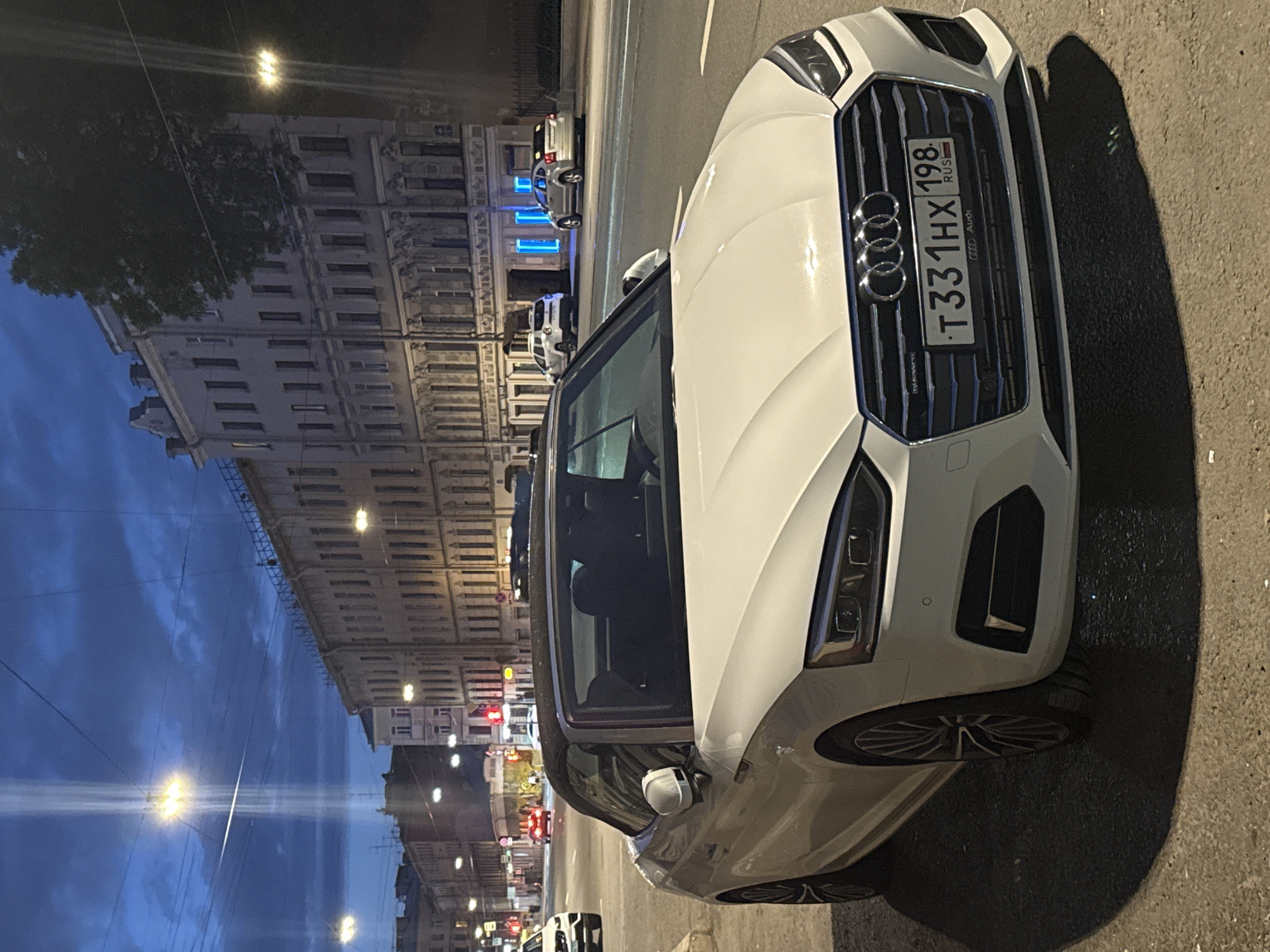 In the city of Saint Petersburg, Russia, a white Audi convertible with a black soft top is parked on the right side of the Nevsky Prospekt, a major thoroughfare. The car is facing the viewer and positioned on a cobblestone sidewalk. Its license plate reads "T331HX 198 RUS". Across the street, a row of old, multi-story buildings with illuminated windows and ornate architectural details line the opposite side of the avenue. The sky is dark blue, indicating dusk or early night, with a single bright light beam piercing through the clouds from a streetlamp. Other vehicles are visible on the street, including a white SUV further down the road and a red vehicle with illuminated taillights. The scene is illuminated by streetlights and the lights from the buildings, casting shadows from the parked Audi onto the pavement.