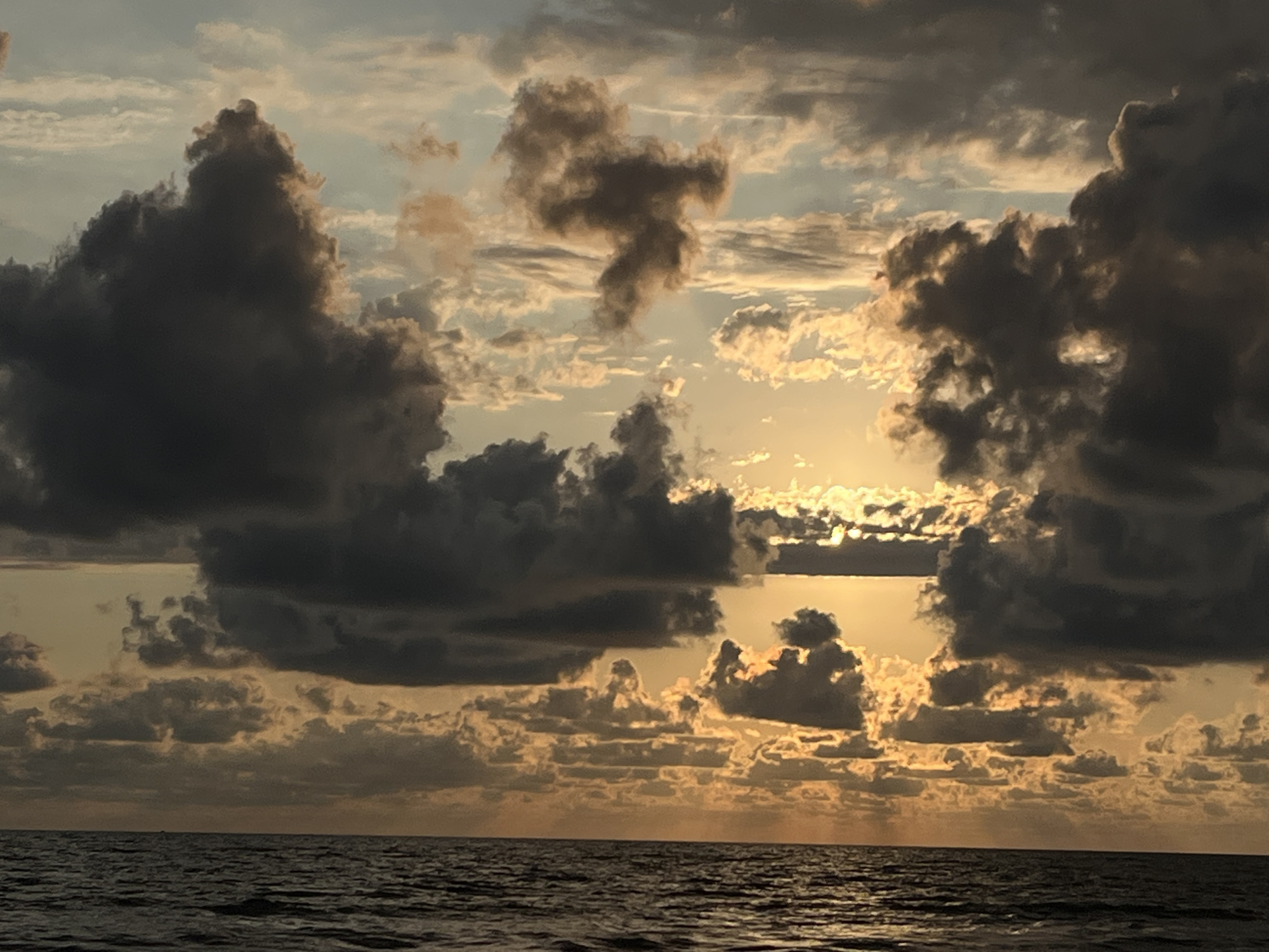 The image shows a dramatic sky filled with dark, heavy clouds interspersed with lighter, illuminated clouds, suggesting a sunset or sunrise over the Black Sea. The sun is visible as a bright, warm glow peeking through the clouds, casting a golden light on the scene. The sea is calm, with gentle ripples reflecting the colors of the sky. The vastness of the sky and the sea creates a sense of awe and tranquility. The photograph is likely taken from a boat or a beach in the village of Supsa, Georgia, a coastal settlement known for its natural beauty and proximity to the Black Sea.