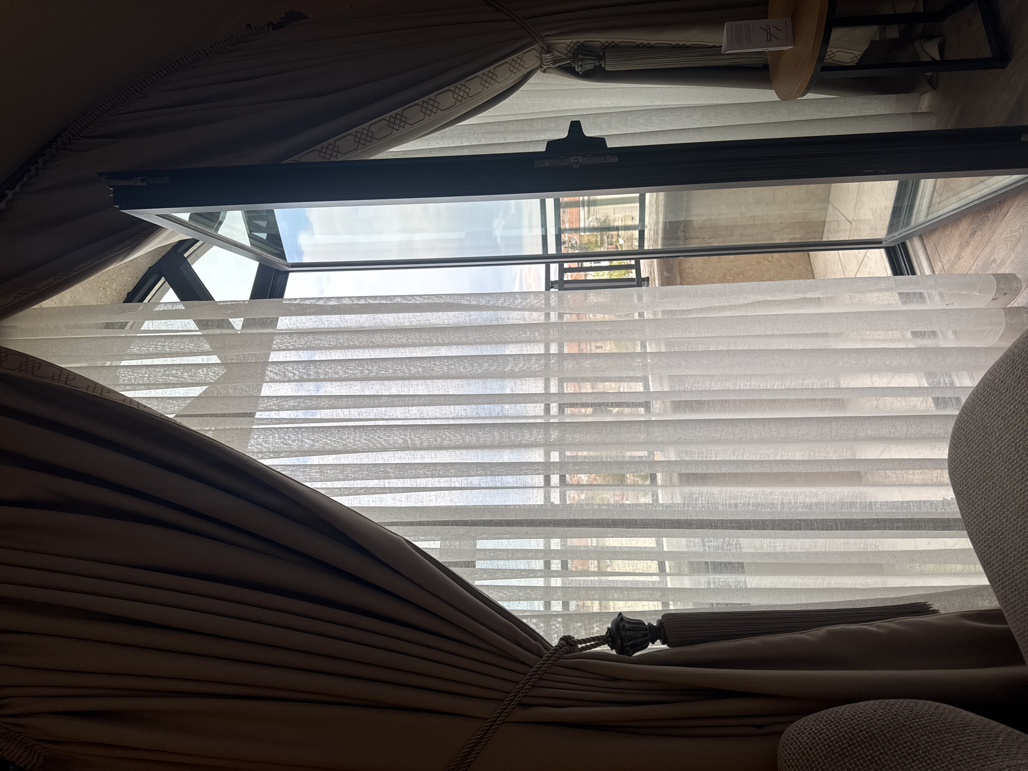 The image shows an interior view looking out through an open window, likely in Ürgüp, Turkey, although specific landmarks are not visible. The window frame is dark grey or black. Sheer white blinds or shades partially obscure the view. Thick brown drapes are drawn to the sides of the window, tied back with a decorative tassel. A small section of what appears to be a chair is visible in the lower right of the frame, covered in a light grey fabric. A small table with a wooden top and metal frame can be seen in the upper right. The view through the window is of a building and sky.