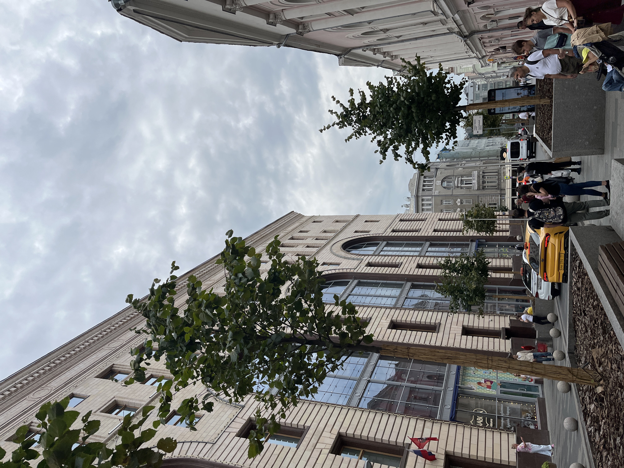 The image depicts a street scene in Moscow, Russia. A large, ornate building with light-colored stone facade and arched windows dominates the left side of the frame. Trees with green foliage line the sidewalk in front of the building, partially obscuring the lower floors which feature large, reflective windows and storefronts. Two flags, one red and one red, white, and blue, are affixed to the building's facade.
On the right side of the street, a narrower, pink building with classical architectural elements, including columns and arched windows, is visible. The street is paved and features a sidewalk with planters and spherical bollards.
Several people are present, some walking along the sidewalk, others gathered in small groups. A yellow taxi is parked on the street, along with a white car. A white van is also visible further down the street. The sky is overcast with a pattern of clouds. The scene appears to be a public urban space, likely a commercial or residential area within the city.