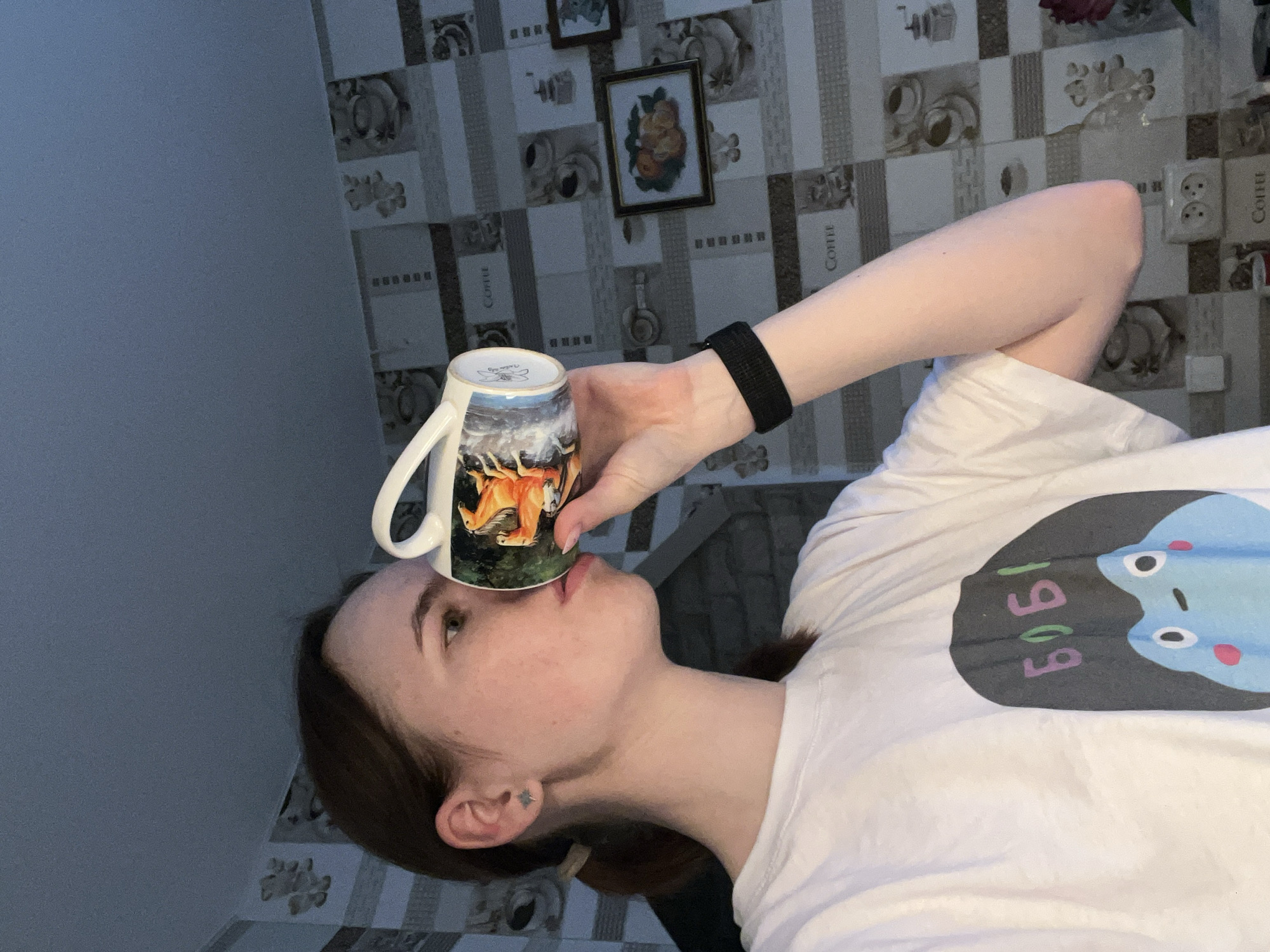 A person is posing with a mug in what appears to be a residential setting. The individual, who is wearing a white t-shirt with a graphic design, holds a mug up to their face. The mug is decorated with a landscape scene. The background is a kitchen with patterned wallpaper adorned with framed pictures. There is no specific evidence in the image to definitively place the location in the city of Chaikovsky, Russia.