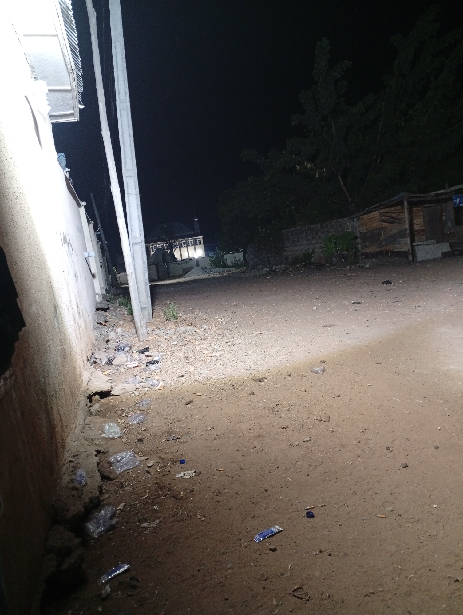Adankolo, Nigeria. Night. A dirt road illuminated by a light source (likely a streetlight or vehicle headlights) runs away from the viewer. The left side of the road is bordered by a wall and several utility poles. Rubbish litters the ground near the wall. On the right side of the road is a structure made of wood and other building materials. A house can be seen far away. No people are visible.