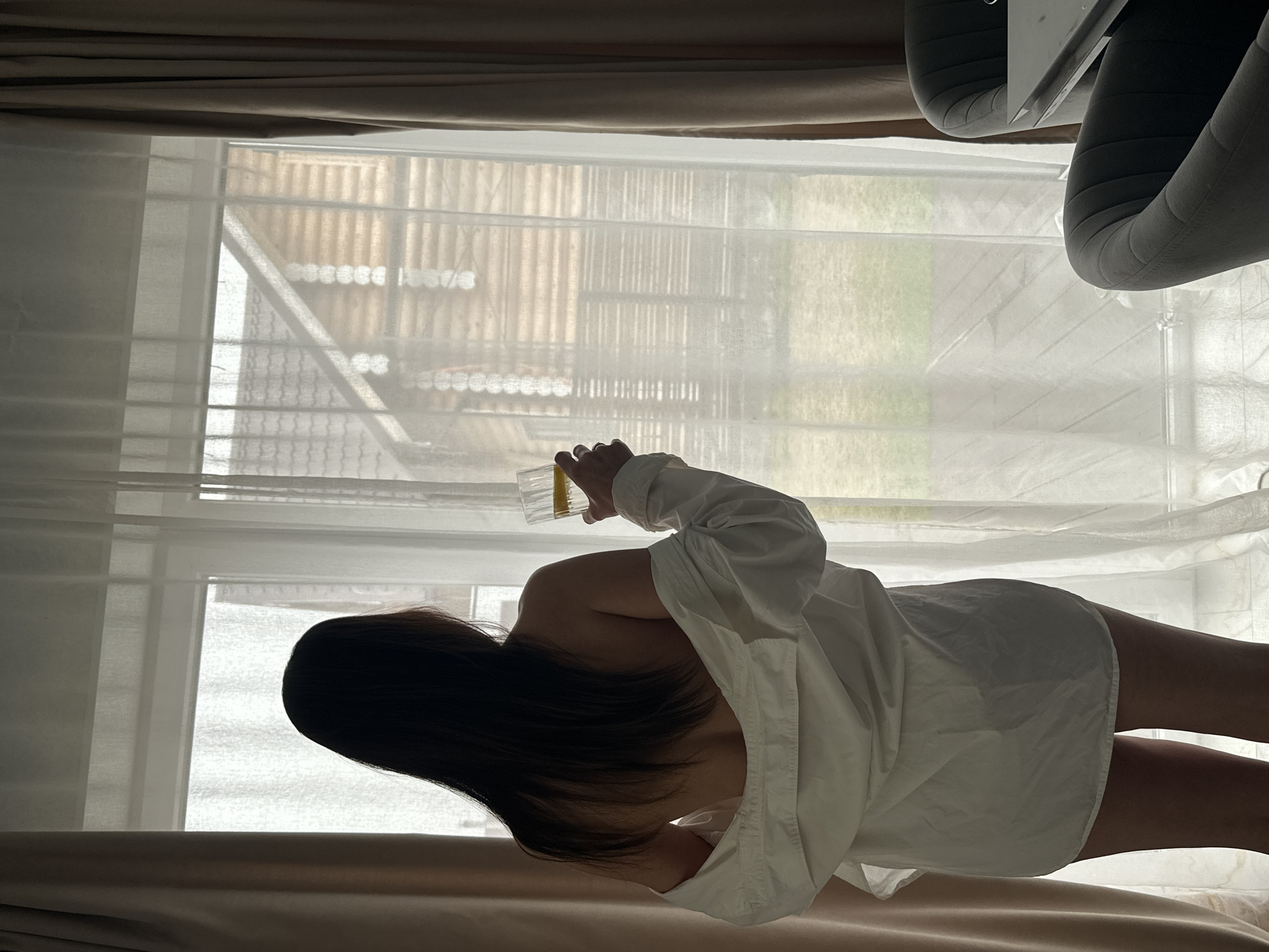 A person, viewed from behind, stands indoors by a window, holding a clear glass containing a yellowish liquid. The individual wears a loose-fitting white shirt, and their long, dark hair cascades over their shoulders. The window is covered by translucent white sheer curtains, which diffuse the natural daylight. Above and below the window frame are opaque brown curtains or blinds.
Through the sheer curtains, an obscured outdoor scene is visible, revealing what appears to be slatted structures, possibly a fence or part of a building, and patches of green land. In the upper left portion of the image, parts of two grey upholstered chairs are visible, suggesting indoor furniture. The overall setting conveys a calm, daytime indoor environment. No text is visible in the image. The location context is Chaikovsky, Russia.