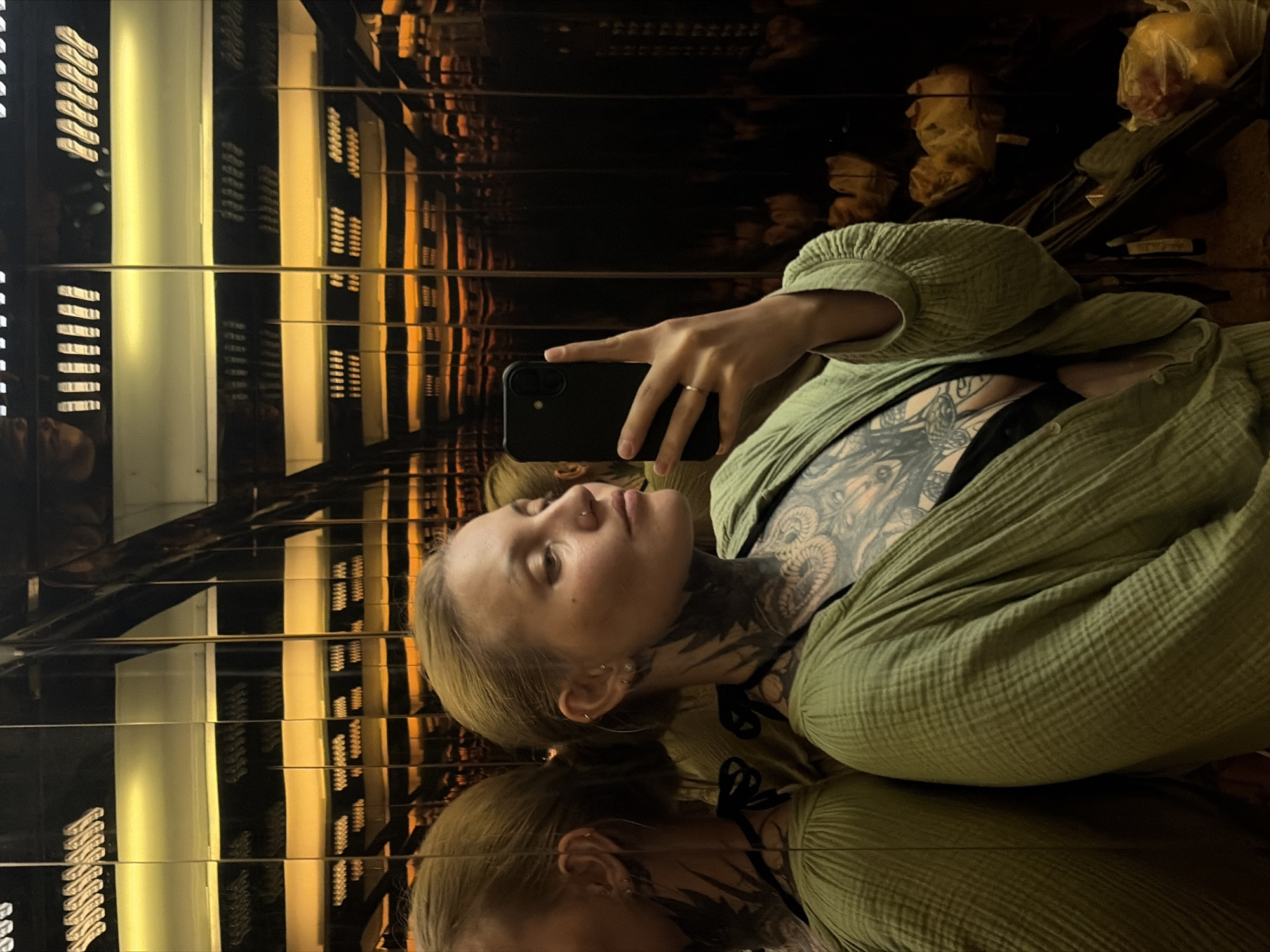 The media file captures a mirror selfie of a person in an indoor setting.
**Subjects:** The primary subject is a young person, appearing female, with fair hair, looking upwards towards her phone. She has extensive black ink tattoos covering her chest, collarbone, and neck, featuring intricate designs including what appears to be a butterfly. She wears multiple piercings in her ears and a small nose piercing. Her reflection is prominently visible below her in the image.
**Objects:** She holds a black smartphone in her right hand, with a simple gold ring visible on her finger. She is dressed in a loose-fitting, textured, olive-green long-sleeved shirt or jacket, worn open over a black top or bra. In the background, indistinct objects resembling plastic bags, possibly containing fruit, are visible in the reflections.
**Scene/Setting:** The setting is an indoor space characterized by highly reflective, dark surfaces on the walls and possibly the ceiling. These surfaces create an illusion of infinite reflections, multiplying linear light fixtures that emit a warm, golden-yellow glow. The environment appears to be a corridor, elevator, or a room designed with mirrored panels, creating a sense of depth and repetition. The location context provided is Đà Nẵng, Vietnam.
**Activity or event taking place:** The person is taking a mirror selfie, actively looking towards her phone to compose the shot.
**Notable details:** The unique reflective surfaces create a visually engaging and somewhat disorienting effect, amplifying the artificial lighting and background elements. The intricate tattoos and the subtle details of her piercings and ring are clearly visible. The overall mood is casual and personal, typical of a selfie. There is no visible text in the image.