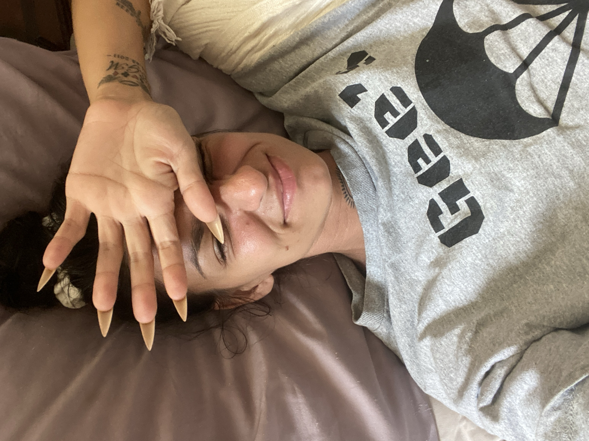 A young woman is lying on a bed, covering her eyes with her hand. She is wearing a grey sweatshirt with a black graphic of a parachute and text that reads "GEE". Her fingernails are long and light-colored. She has dark hair pulled up in a bun. A tattoo is visible on her wrist. The background is a purple sheet. The scene is indoors. There is no indication of the location being in Mũi Né, Vietnam.
