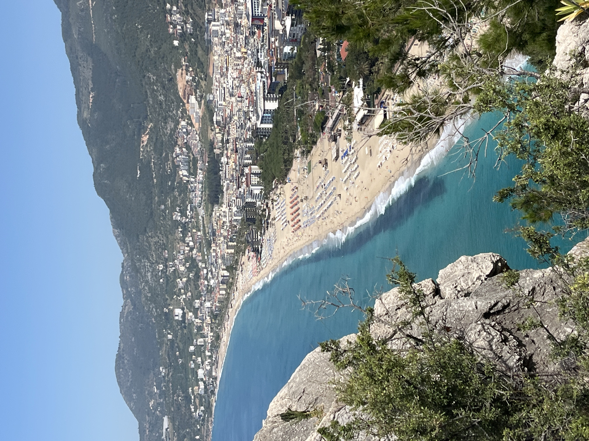 An eye-level shot from a high vantage point shows the city of Alanya, Turkey, and its beach from a slightly turned right angle. The sky is a clear blue. A tall, sloping mountain runs vertically on the left side of the image. The city buildings are located near the base of the mountain, and there is a large beach located between the city and the bright blue water. The beach is filled with many beach umbrellas and some people sunbathing. White wave crests are visible in the water. On the right side of the image are some rocks and green foliage.