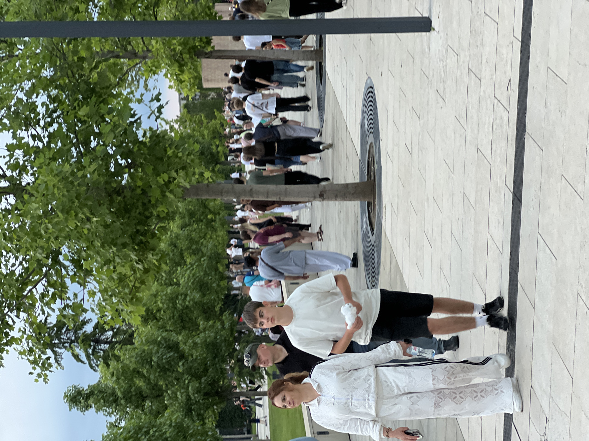 The image depicts a group of people walking along a paved pedestrian walkway lined with trees. In the foreground, a woman in white patterned tracksuit and a teenage boy in a white t-shirt and black shorts are walking towards the viewer. The woman is holding a phone in her left hand and the boy is holding a white object in his hands. Behind them, several other individuals are visible walking in the same direction, some in casual attire. The environment appears to be a park or a public square with trees and a light-colored paved surface. The background shows more trees and distant buildings. The scene suggests a casual outdoor activity or public gathering. The image was taken in Krasnodar, Russia, at the "Gallery" shopping mall, specifically on the pedestrian zone in front of it.