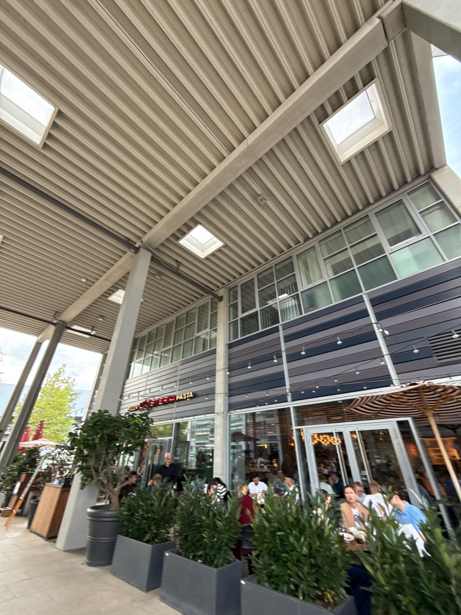 The image shows the exterior of a restaurant in Wolfsburg, Germany, with a prominent sign that reads "PIZZA & PASTA". The restaurant has a modern architectural design with large glass windows and a sloped, corrugated metal roof. Several people are seated at tables outside the restaurant, some under a striped awning. There are also potted plants and trees in planters, adding greenery to the scene. The overall impression is of a busy outdoor dining area on a cloudy day.