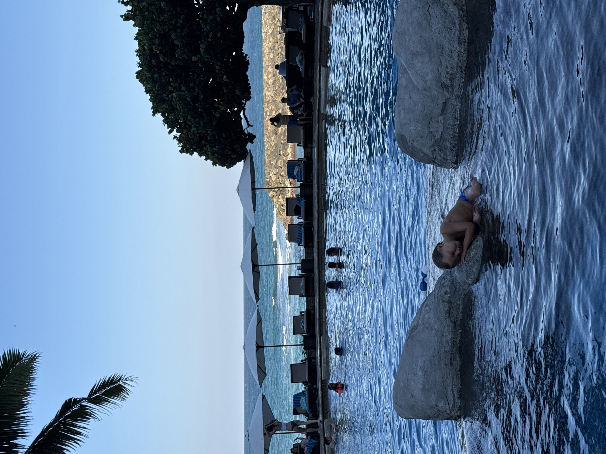 In Kuta Selatan, Indonesia, a young child sleeps on a large rock in a swimming pool. The child is wearing blue swim trunks and is positioned on their side, with their head resting on the rock. The pool is filled with blue water, and there are other rocks visible in the foreground. In the background, there is a row of lounge chairs with umbrellas, a stone wall, and the ocean. Several people are visible in the pool and sitting on the lounge chairs. Palm fronds hang from the upper left of the frame, and a large tree with dark green foliage is to the right. The sky is clear and blue.