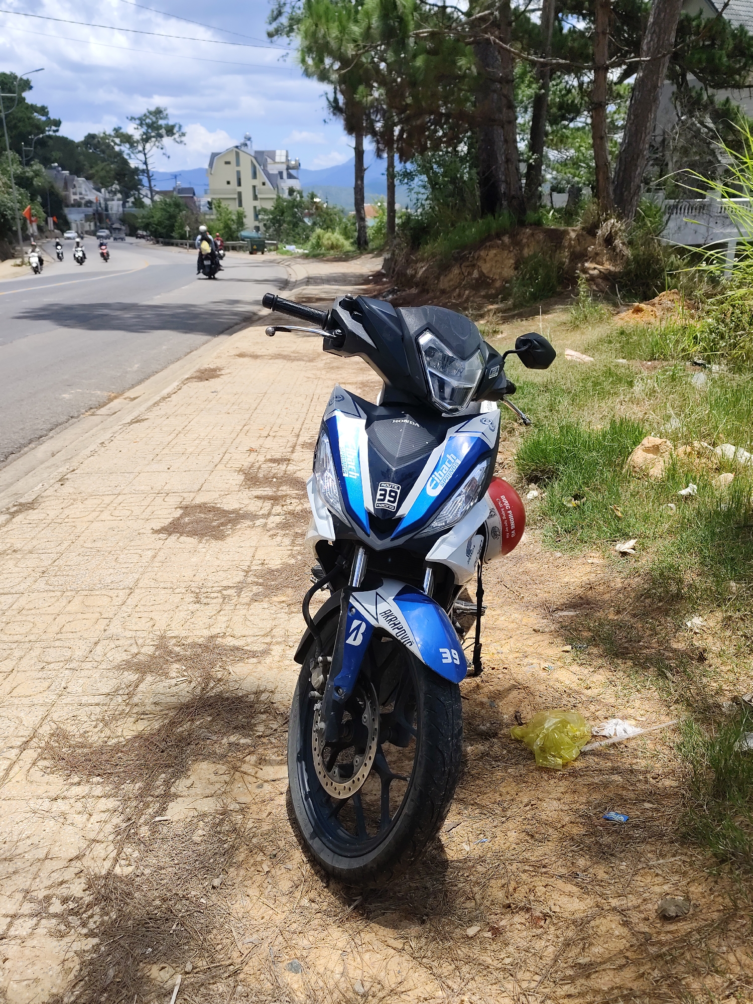 The image features a Honda motorcycle parked on the side of a road in the city of Xuân Trường - Đà Lạt, Vietnam. The motorcycle is predominantly white and blue with visible stickers and markings. The front tire of the motorcycle is turned slightly to the left.
The motorcycle is parked on a paved area of the road, which is patterned with square segments. Next to the paved area is a section of dirt and grass. A discarded yellow plastic bag is visible near the front tire of the motorcycle.
In the background, the road curves away with other motorcycles traveling on it. Further back, buildings and mountains are visible under a cloudy sky. Trees are present along the roadside, contributing to the landscape of the city.