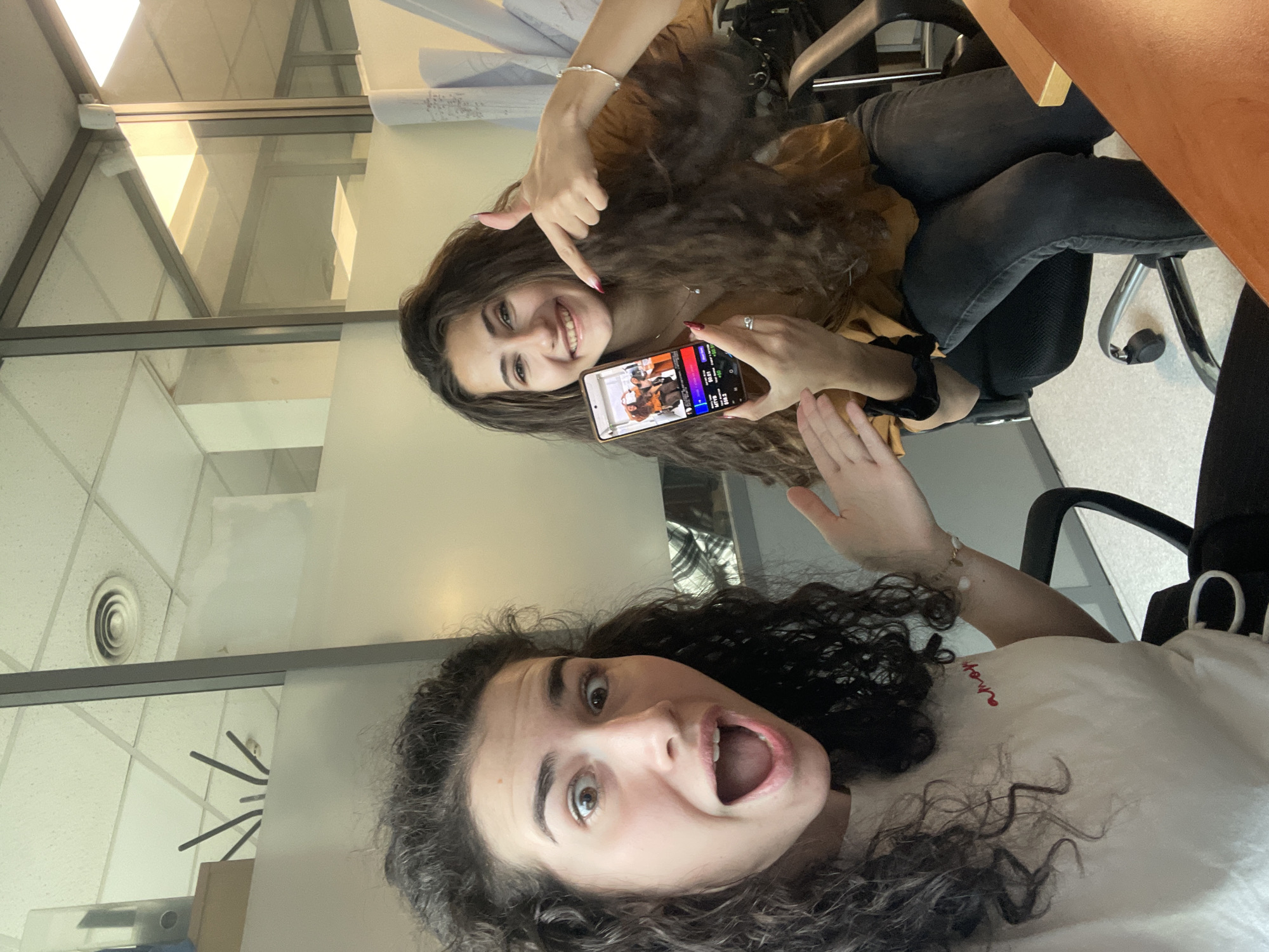 Two young women are in an office setting. One woman is seated and holding a mobile phone displaying an image of both women. She is pointing at the image on the screen and smiling. The other woman is in the foreground, her mouth open in a surprised expression. The office environment includes a window, fluorescent lighting, ceiling tiles, office chairs, and a desk. The location is likely in Sofia, Bulgaria.
