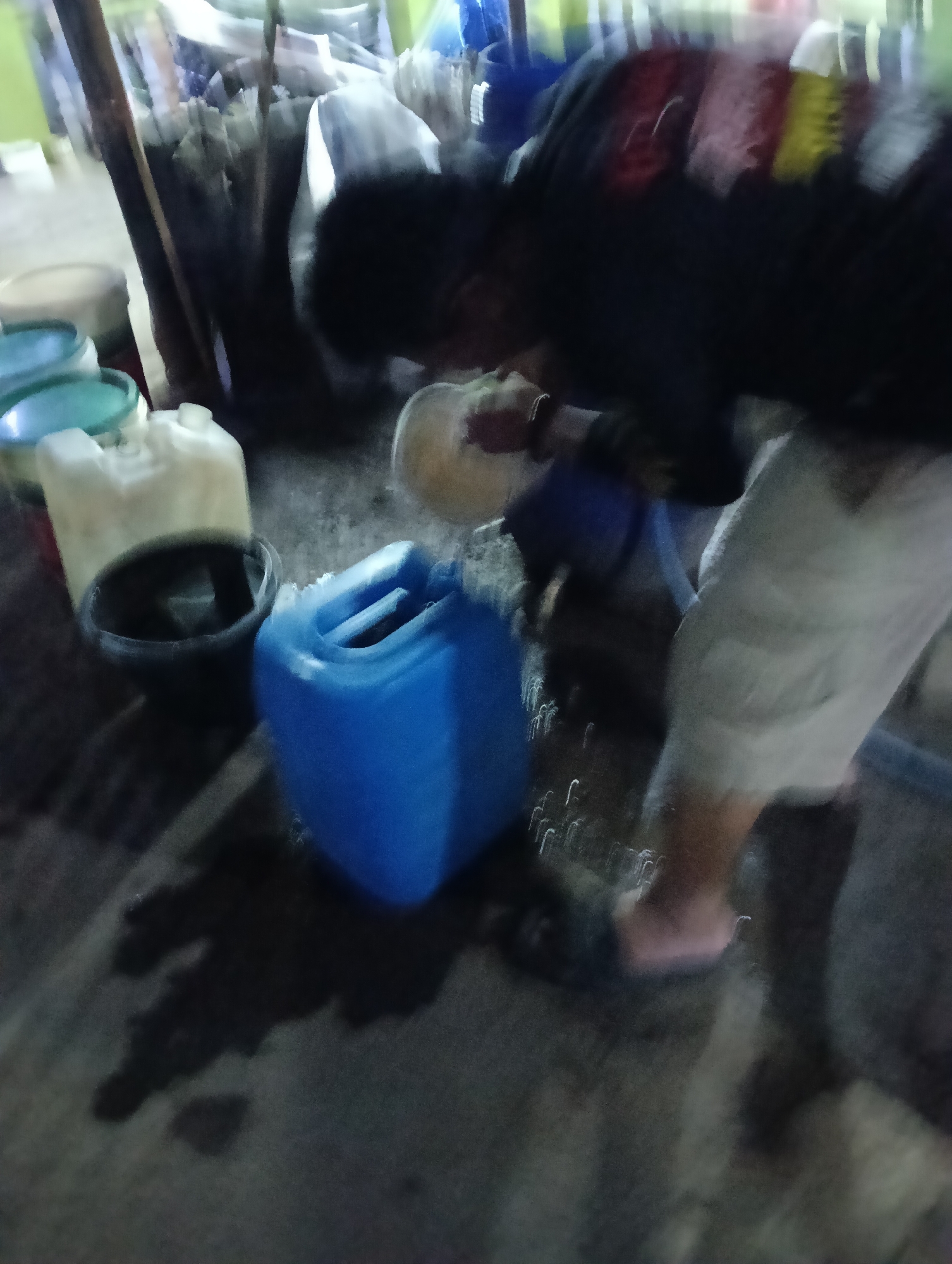 The image captures a blurry scene of a person, likely a male, leaning over to interact with several large plastic containers. The environment appears to be outdoors, possibly a roadside or an open-air market, given the presence of multiple containers and what looks like debris or spilled liquid on the ground. The lighting is dim, suggesting it might be evening or night.
In the foreground, a large blue plastic container is prominent, with its handle clearly visible. To its left, a pale yellow container, also plastic, sits partially filled with a dark liquid. A black bowl or bucket is placed next to this yellow container. Further to the left, there are other containers with blue and green lids, though their details are less clear due to motion blur.
The person is wearing a dark shirt and light-colored pants. Their upper body is blurred, indicating movement, and they appear to be reaching towards or pouring something from a circular object held in their hands, possibly a ladle or a smaller container, into one of the larger containers.
The ground is a dark, uneven surface with some lighter patches that could be reflections or wet areas. The background is indistinct, with blurred shapes that might represent structures or foliage, and streaks of light suggesting artificial illumination or movement.
This scene could be typical of a water vendor or someone refilling or distributing liquids from bulk containers in a less developed area. Without further context, it's difficult to pinpoint an exact location within Tasikmalaya, Indonesia, but it aligns with scenes that might be found in informal commercial areas or residential neighborhoods where bulk water is sold.
