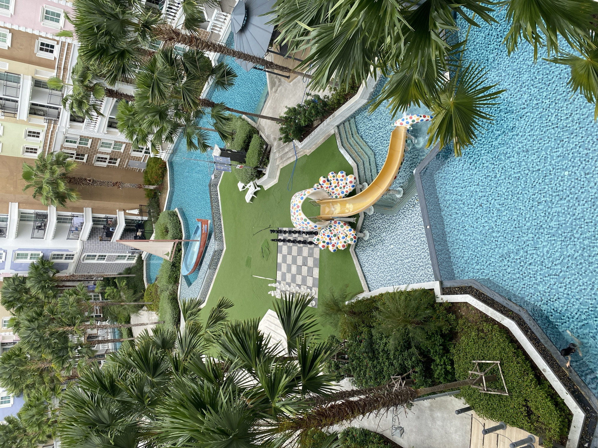 An aerial shot captures a resort in Pattaya City, Thailand, featuring a large swimming pool with turquoise, tiled water. Palm trees and lush greenery surround the pool area. A playful yellow slide with a colorful, polka-dotted base is positioned within the pool complex. Adjacent to the pool is a green lawn area that hosts an oversized chessboard with white and black pieces. A decorative red boat with a sail is situated in a smaller, shallow pool. In the background, multiple resort buildings with balconies and pastel-colored facades are visible, interspersed with more palm trees. The image also shows a shaded gazebo and several white lounge chairs. A single person is visible swimming in the main pool.