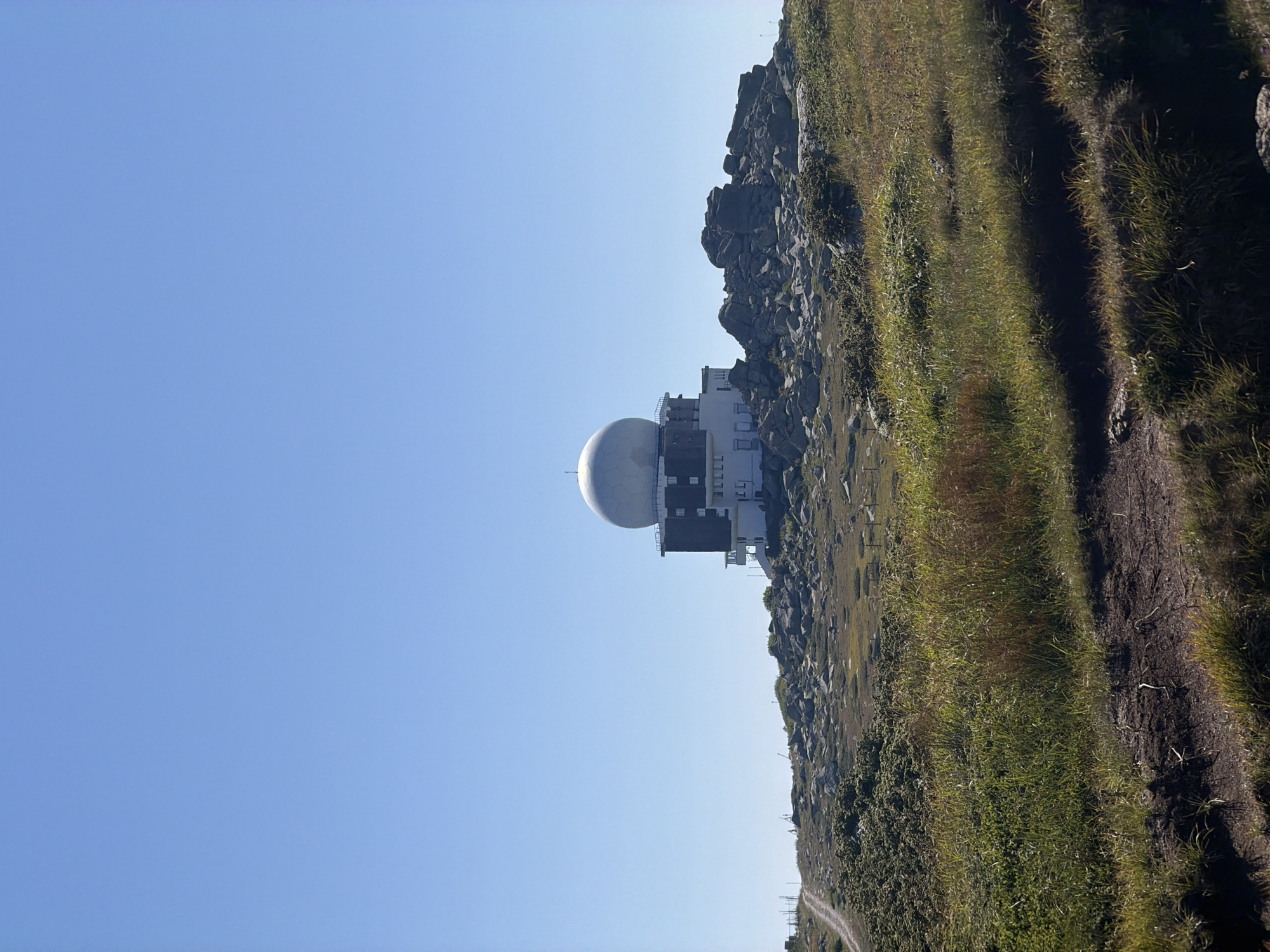 An outdoor, low-angle, full shot shows a large, white, spherical dome perched atop a dark, multi-story building with numerous windows, which is situated on a rocky, grassy hillside. The building is on the right side of the image, against a clear, bright blue sky. A dirt road or path winds up the left side of the hillside. The landscape is rugged and appears to be mountainous, with scattered rocks and sparse vegetation. The sky is a dominant feature, taking up approximately the top half of the frame. The image is dated August 14, 2023, and is described as being taken in Bistritsa, Bulgaria, which is the location of the Bulgarian Antarctic Base.