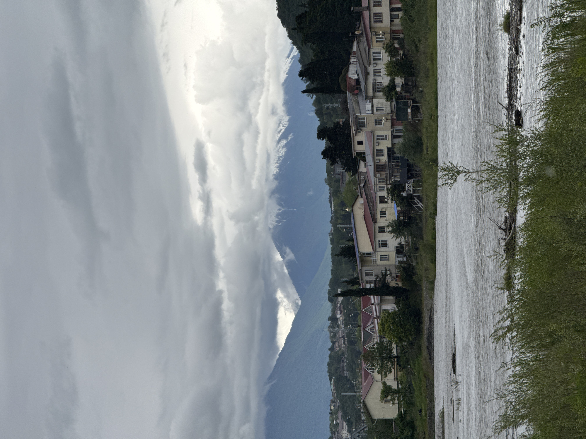 The image depicts a landscape in Sochi, Russia, featuring a mountain range shrouded in clouds on the left, a river in the center, and residential buildings on the right. The sky is overcast with heavy clouds. The mountain range is partially obscured by the clouds, with some snow visible on the peaks. The river is flowing rapidly and appears turbulent. The residential buildings are multi-storied and situated along the riverbank. Trees and bushes are present along the river's edge.