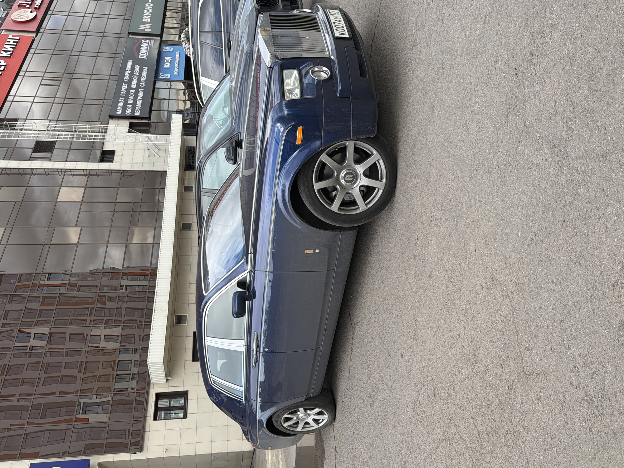 A navy blue Rolls Royce with license plate K007AY777 is parked next to a building with signs in Russian. The signs advertise "Laminate Parquet Quartz Vinyl," "Wallpaper Paint Molded Decor," "Ceramogrant Sanitary Ware," and "Domix," "Vkusno", "MegaFolis," "Vhod - Welcome," "Burger King" with logo and store hours. The Rolls Royce is parked on a gray asphalt surface. The building has a modern design with reflective glass windows. The scene appears to be in Moscow, Russia based on the license plate and the Cyrillic lettering on the signs.