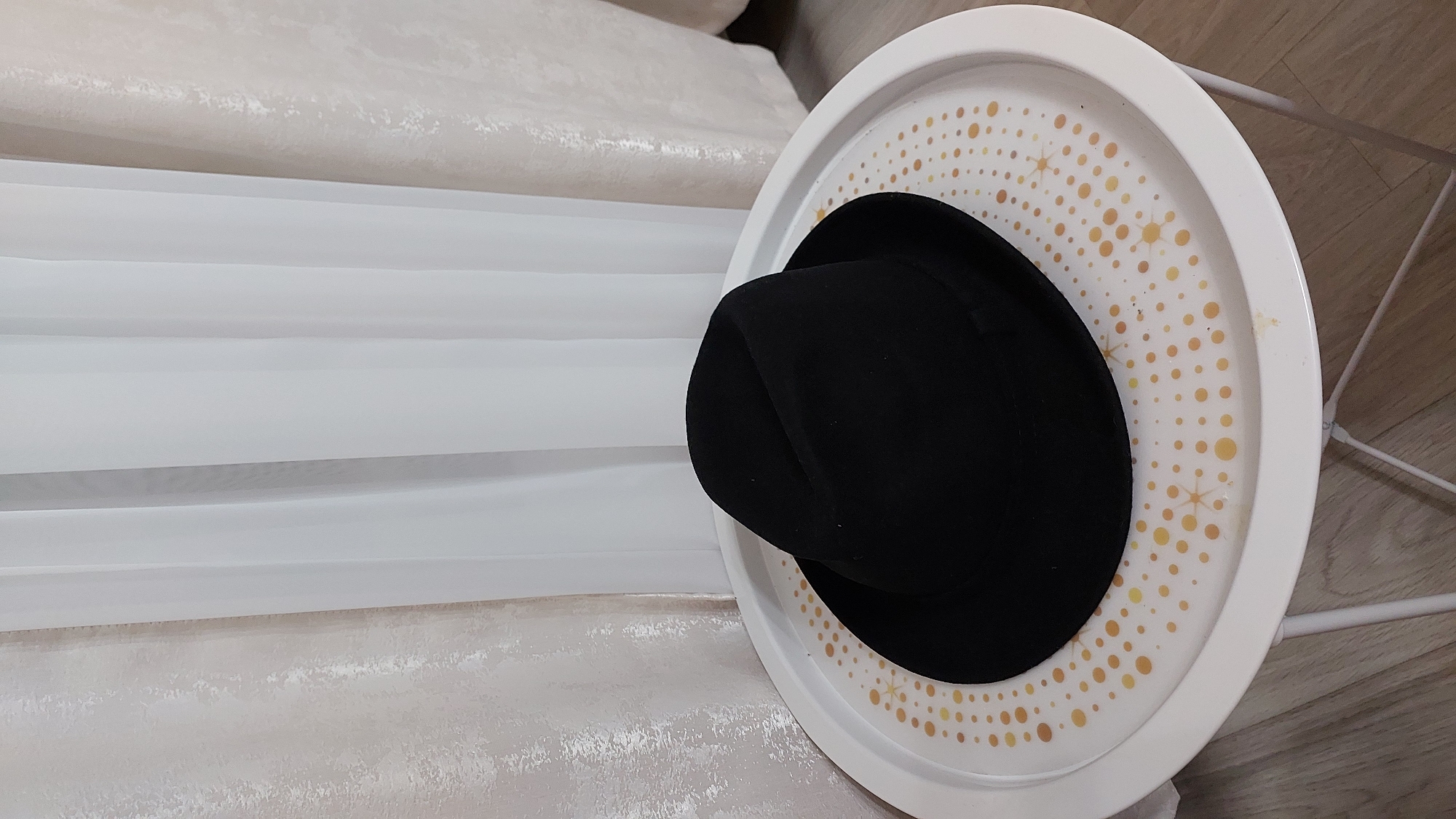 A black fedora hat is centrally placed on a white, circular side table with a decorative pattern of small orange and yellow dots. The table has thin white legs and is situated on a light brown wooden floor. In the background, sheer white curtains with a subtle textured pattern are visible, suggesting an indoor setting. The lighting is soft and diffused. There is no indication of the city of Novocheboksarsk, Russia.