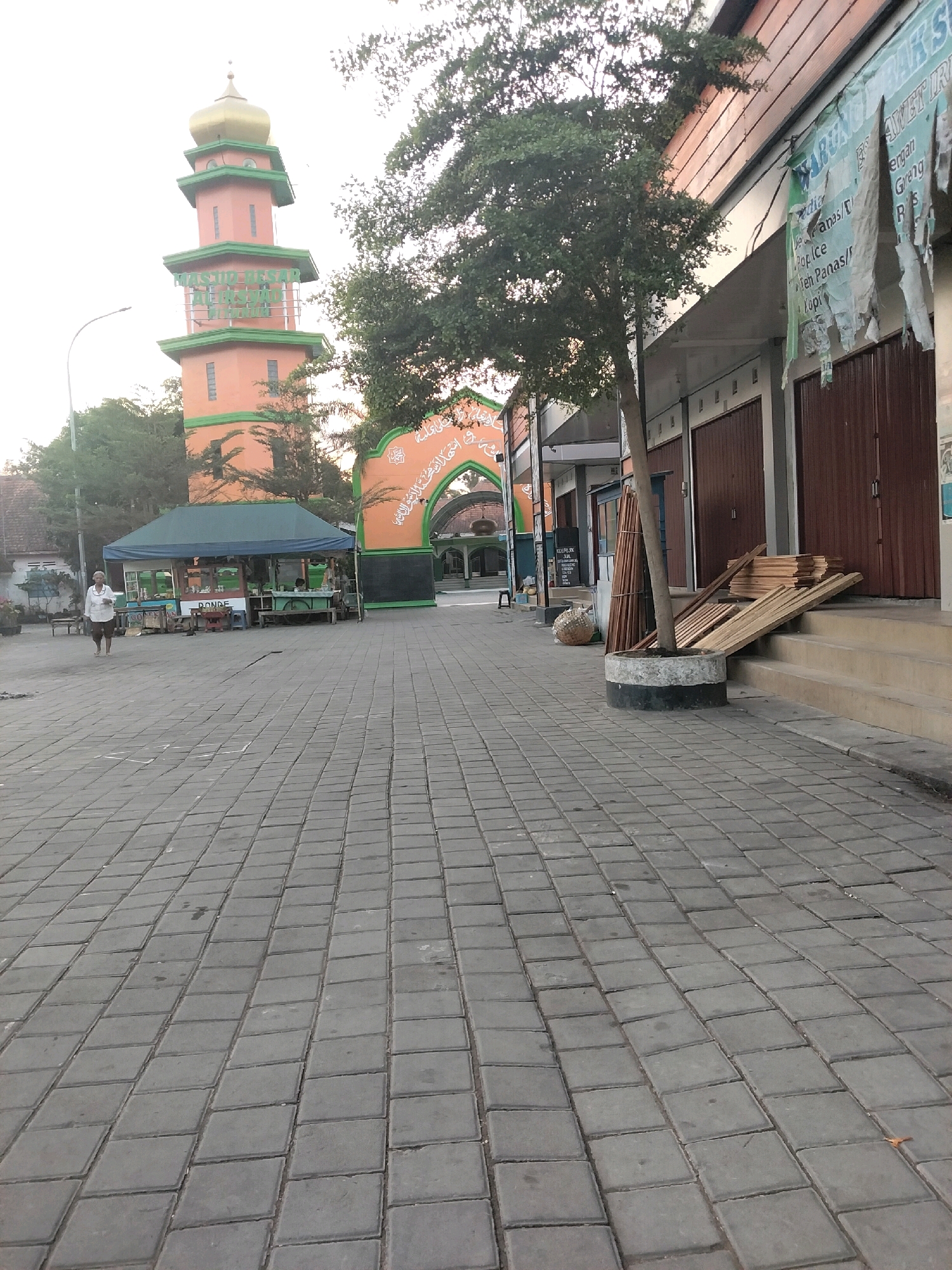 The image captures a view of a mosque minaret and surrounding buildings in Pituruh, Indonesia. The minaret, painted orange with green accents and a golden dome, stands prominently. In front of the mosque's entrance archway, a small food stall with a green canopy is visible, with a person standing nearby. To the right of the scene, a row of shops with wooden shutters lines the paved walkway. Stacked lumber is placed on the steps leading up to one of the shops. The ground is covered with gray interlocking pavers. The overall lighting suggests it is either early morning or late afternoon.