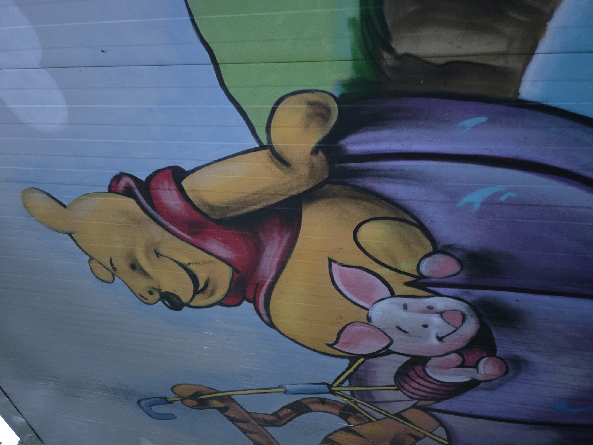 This image is a mural depicting Winnie the Pooh, Piglet, and Tigger. Pooh, wearing a red shirt, appears to be airborne. Piglet is close to Pooh, seemingly being carried along. Below them is Tigger, holding onto some sort of implement, which connects to the other characters. The setting is an outdoor urban environment in the city of Unknown, Unknown. The mural is painted on a surface that resembles wooden planks.
