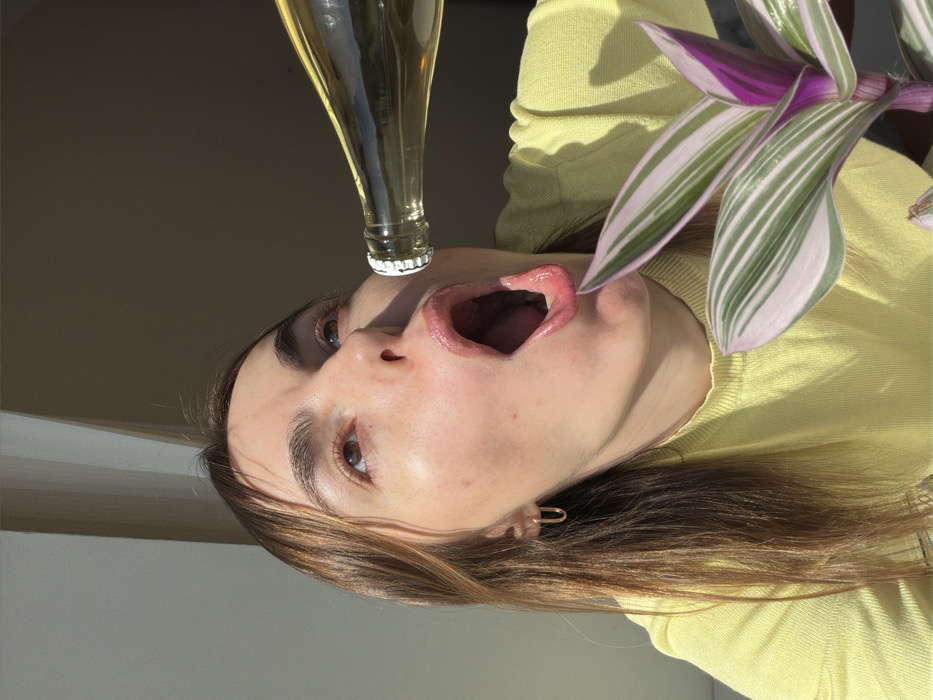 One female subject with medium-length brown hair, wearing a light yellow long-sleeved garment and a gold-colored U-shaped hoop earring in her left ear, is depicted in an indoor setting. Her head is tilted back, eyes gazing upwards, and her mouth is wide open, revealing the oral cavity and a slightly curled tongue. A clear glass bottle, sealed with a white crown cap and containing a pale yellow liquid, is positioned horizontally in front of her open mouth, with its opening directed towards her. In the lower right foreground, a potted plant with variegated green and pink/purple leaves is visible. Strong, direct sunlight illuminates the scene from the left, casting shadows on the plain, light-colored interior wall in the background. The specific location within Izhevsk, Russia, cannot be determined from the visual information provided.