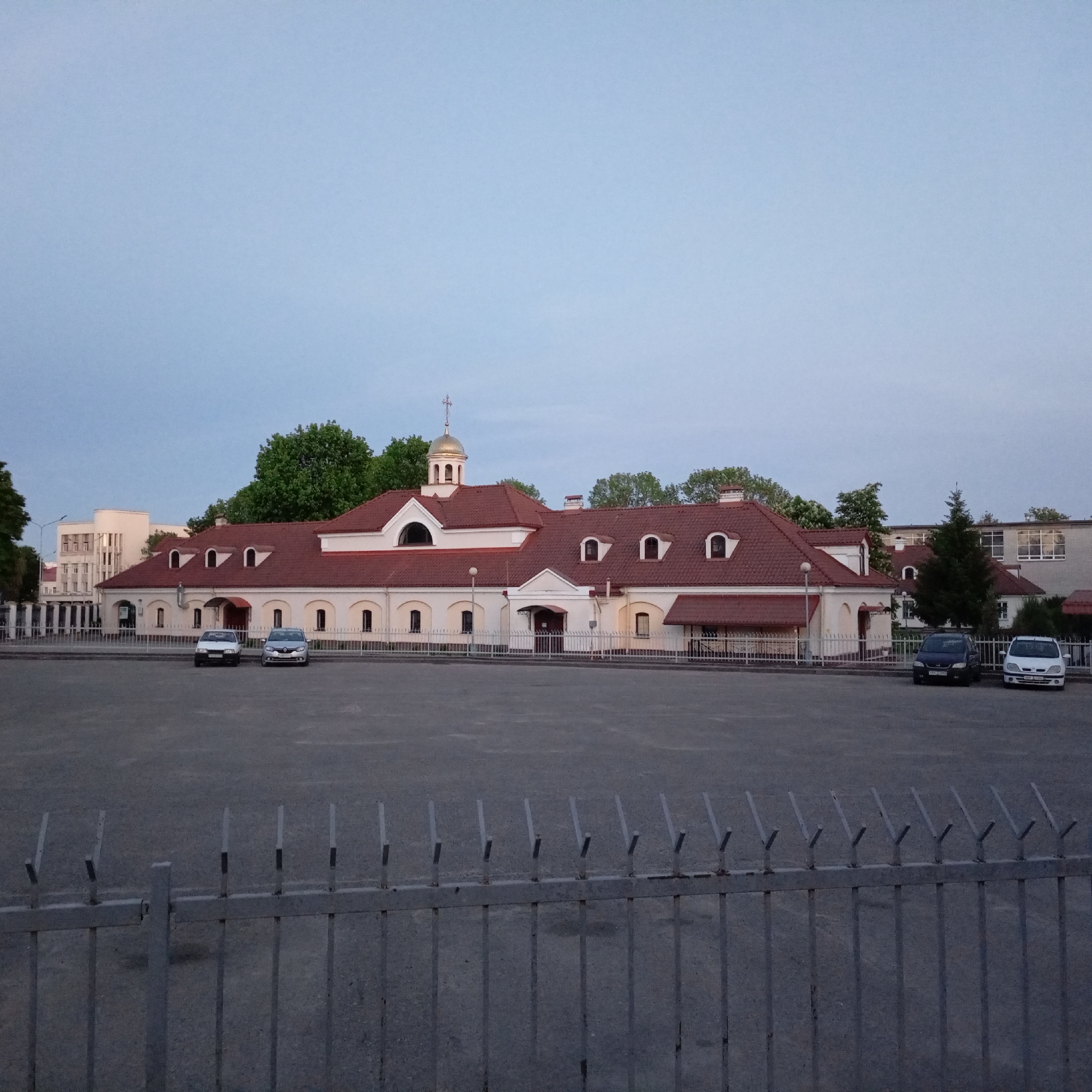 The image depicts a building in Hrodna, Belarus, likely a church or religious structure, judging by the presence of a golden dome topped with a cross. The building is predominantly white with a red roof. The structure has a long, rectangular shape with a prominent entrance in the center and dormer windows along the roofline. In front of the building is a large, empty parking area or plaza. Several vehicles are parked in the lot. A metal fence runs along the bottom edge of the frame, separating the viewer from the parking area. The sky is a clear, light blue, suggesting either early morning or late afternoon.