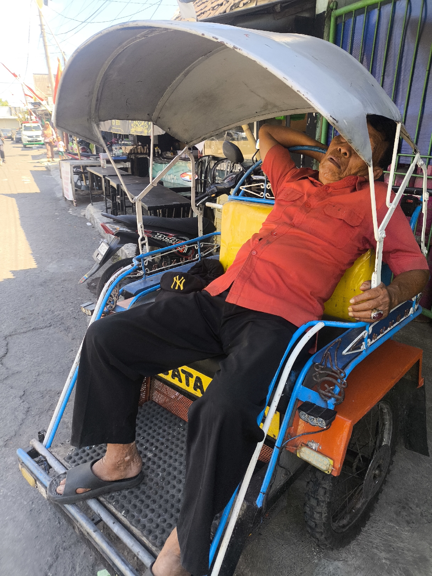 An elderly man in a red collared shirt and black pants is asleep in a three-wheeled vehicle, possibly a becak or pedicab, on a street in Lumajang, Indonesia. The vehicle has a large, gray canopy overhead and a yellow seat. The man is lying back with his right arm behind his head and his mouth slightly open. A black baseball cap with a yellow "NY" logo is visible on the seat. The street appears to be paved, with some cracks and wear. In the background, there are several motorcycles parked and a row of tables and chairs, suggesting a market or food stall area. Other people and vehicles are visible in the distance. The lighting suggests it is daytime.