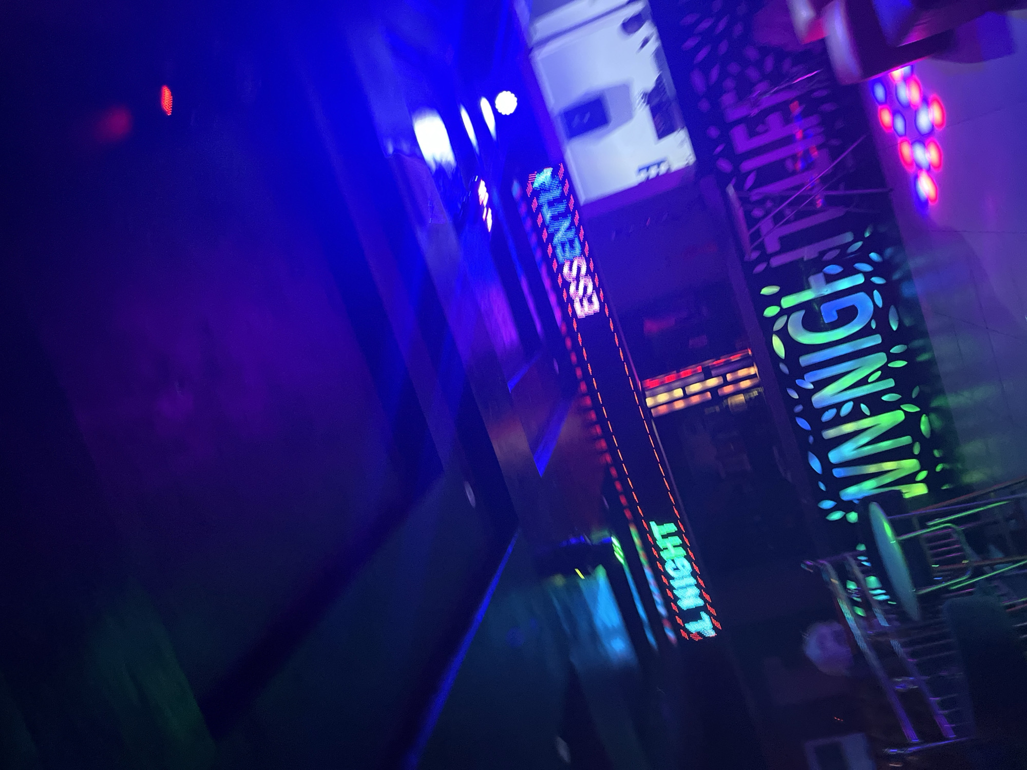 The image depicts an indoor scene illuminated by vibrant neon lights. The environment appears to be a club or entertainment venue. In the foreground, a sign with green neon lettering reads "NN NIGHT LIFE" with decorative leaf patterns surrounding the text. To the left of this sign, a table and chairs are visible, suggesting a seating area. Above the scene, a horizontal sign with red and green lights displays the word "NIGHT". Further to the right and higher up, another sign with blue and green neon lights spells out "ESSENTIAL". The ceiling is dark, with streaks of green and blue light suggesting overhead lighting or projections. The overall ambiance is dark and energetic, characteristic of a nightlife establishment. There are no identifiable people in the image, and the scene is captured at an angle, giving a dynamic perspective. The location is stated to be Unknown, Unknown.