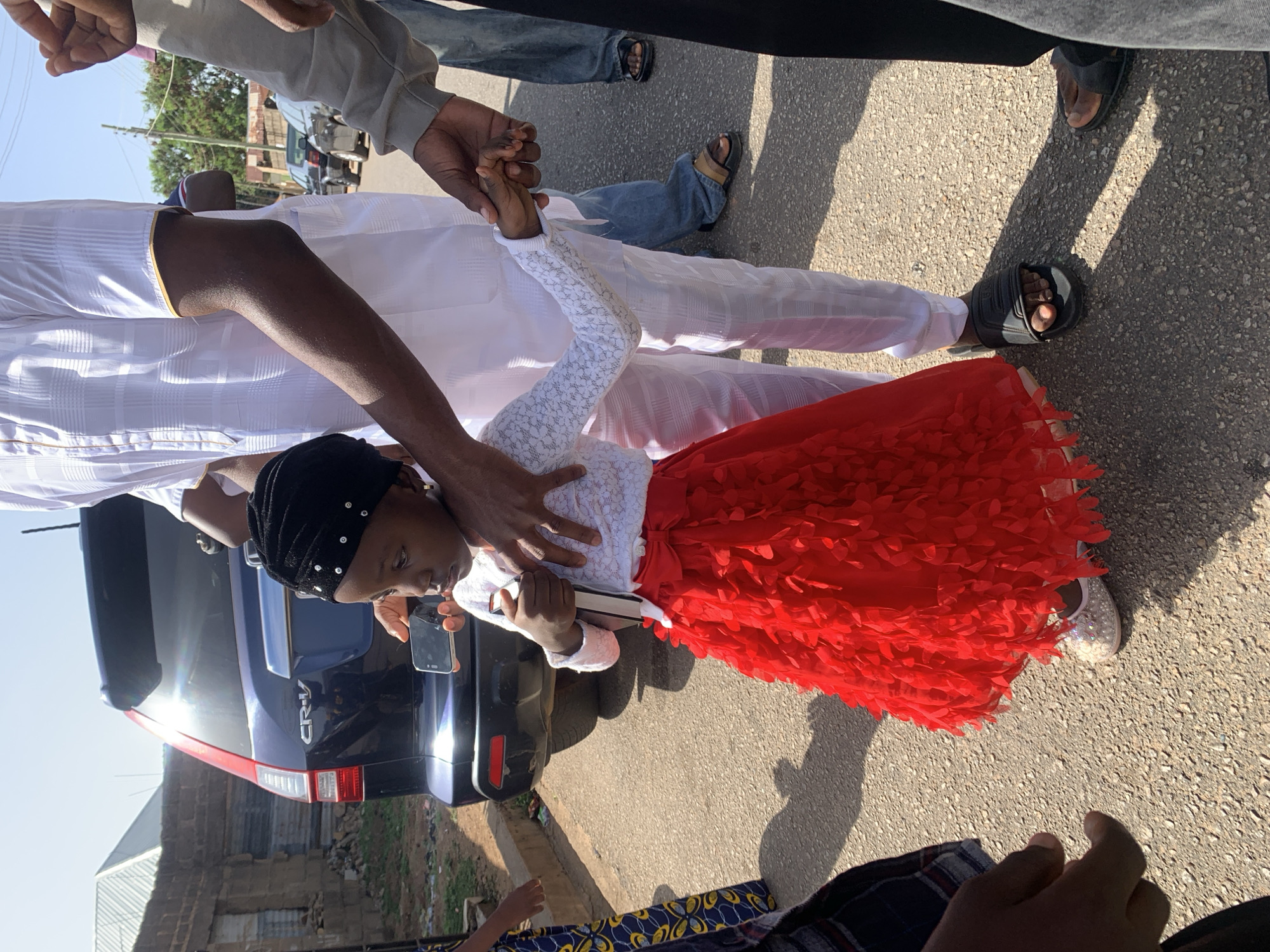 A young girl in a white lace top and red dress stands on a paved road, appearing to be supported by an adult male. The man is wearing a white outfit, commonly seen in the city of Jos, Nigeria. The girl is holding a book and looking down, while the man has his hand on her shoulder. Behind them is a dark blue Honda CR-V. The background shows a building and other individuals, indicating an urban setting. The lighting suggests it is daytime.