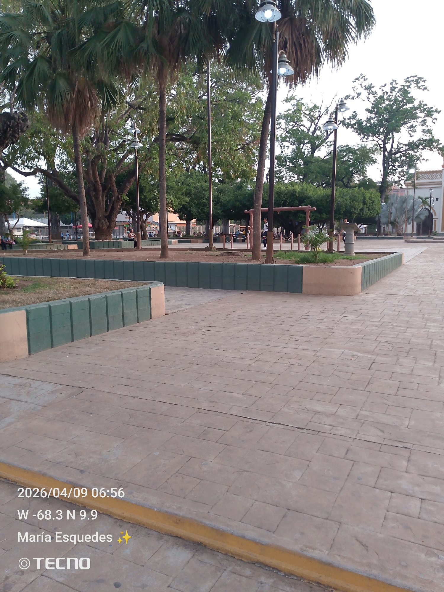 The image shows a public plaza in Tinaquillo, Venezuela on April 9, 2026, at 6:56 AM. The location is approximately at -68.3° West and 9.9° North, and is credited to María Esquedas, using a Tecno brand camera. The plaza features a paved area with a pattern resembling large stone blocks. There are elevated planters with green and tan borders containing grass and small plants. Several trees, including palm trees, are visible. Street lamps are positioned throughout the plaza. In the background, there are people walking and seated, and a playground structure can be seen. Buildings are visible in the far background.