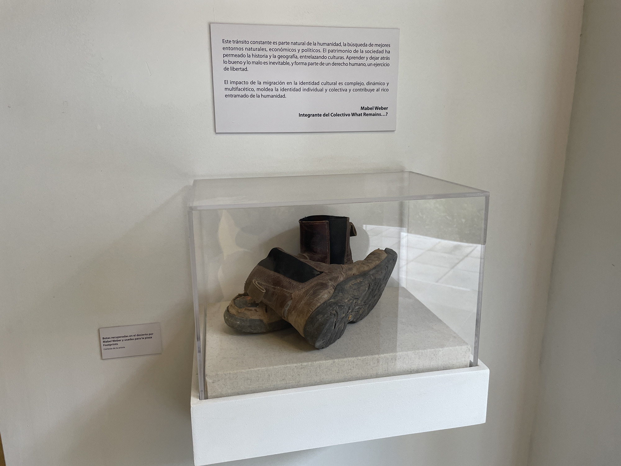 The image shows an art installation on display. There are two signs on a white wall. The first sign contains text in Spanish, and reads "Este transito constante es parte natural de la humanidad...", ending with "Integrante del Colectivo What Remains...?". The second sign is smaller and located lower to the left. It reads "Botas recuperadas en el desierto por Mabel Weber y usadas para la pieza Footprints Cortesia de la artista". Below these signs is a clear acrylic display case. Inside the case are a pair of worn, brown leather boots, set upon a light gray surface. The display case has a white base.