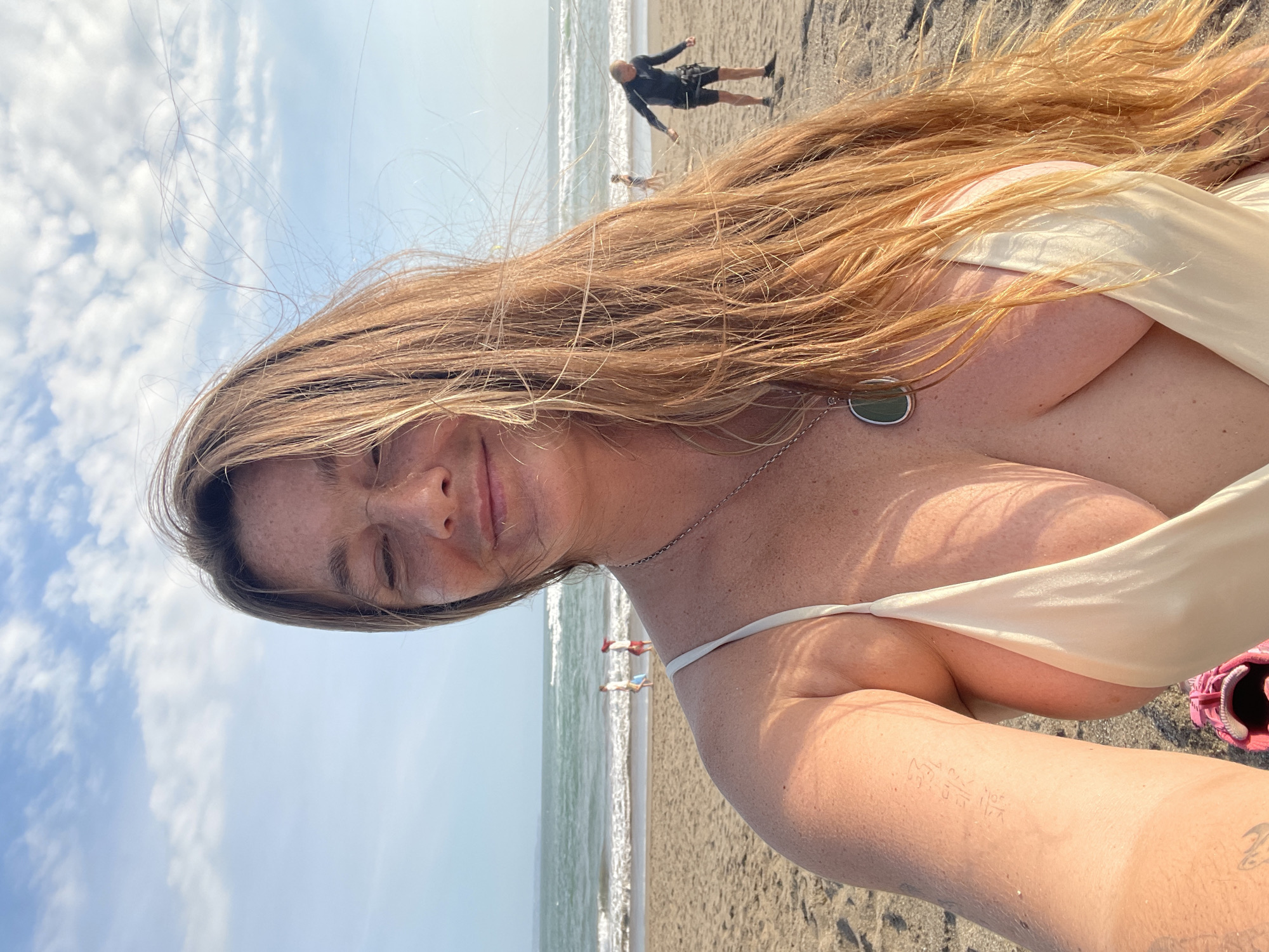 A woman with long, light brown hair and freckles takes a selfie on a beach in North Kuta, Indonesia. She is wearing a light-colored, low-cut bikini top and a necklace with a green pendant. Her eyes are closed and she is smiling. In the background, the ocean is visible with waves breaking on the shore. Several people are walking on the beach in the distance. The sand is light brown, and the sky is blue with scattered clouds. The image appears to be taken during the day, likely in the late afternoon due to the angle of the sun.
