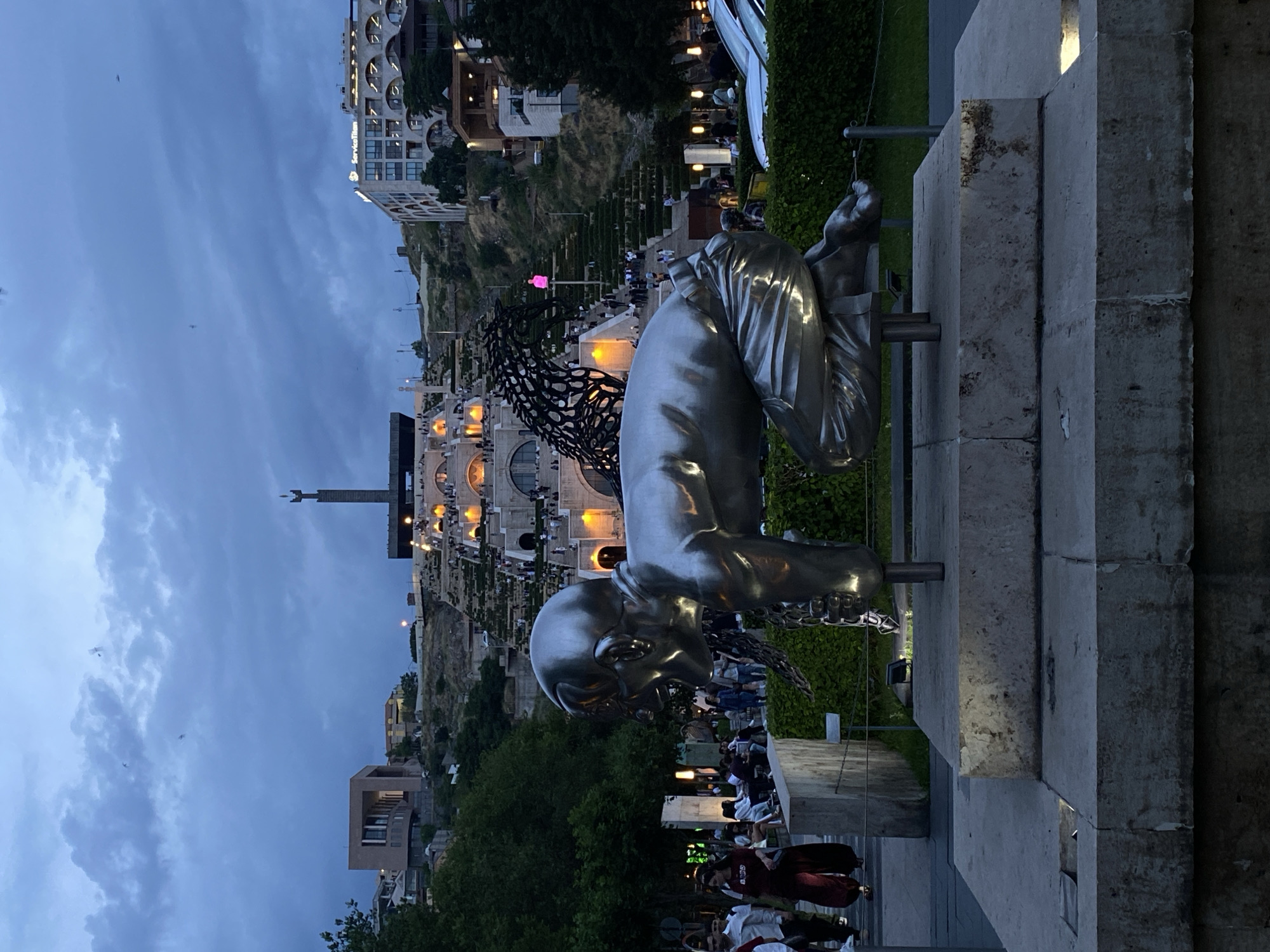 The image captures an urban public space at dusk, featuring prominent art installations and a lively atmosphere. In the foreground, a large, polished metallic sculpture of a man is depicted on all fours, appearing to crawl or reach forward, resting on a stone pedestal. Behind this figure, an abstract, dark-colored sculpture with intricate, organic, web-like patterns is also visible.
The background is dominated by a large, multi-tiered stepped monument, strongly resembling The Cascade in Yerevan, Armenia, which is illuminated by warm lights. Surrounding the monument are various buildings with distinct architectural styles, some also lit up. Numerous people are present throughout the scene; some are strolling on paths, while others are gathered or sitting on the steps of the monument, enjoying the evening.
The sky is a deep twilight blue, partially covered with clouds, indicating the time of day is dusk or early evening. Artificial lights from buildings, streetlights, and the illuminated monument create a warm glow, contrasting with the fading daylight. On a modern building in the upper right quadrant of the image, the illuminated text "ServiceTitan" is visible. The scene portrays a vibrant cultural and social hub.