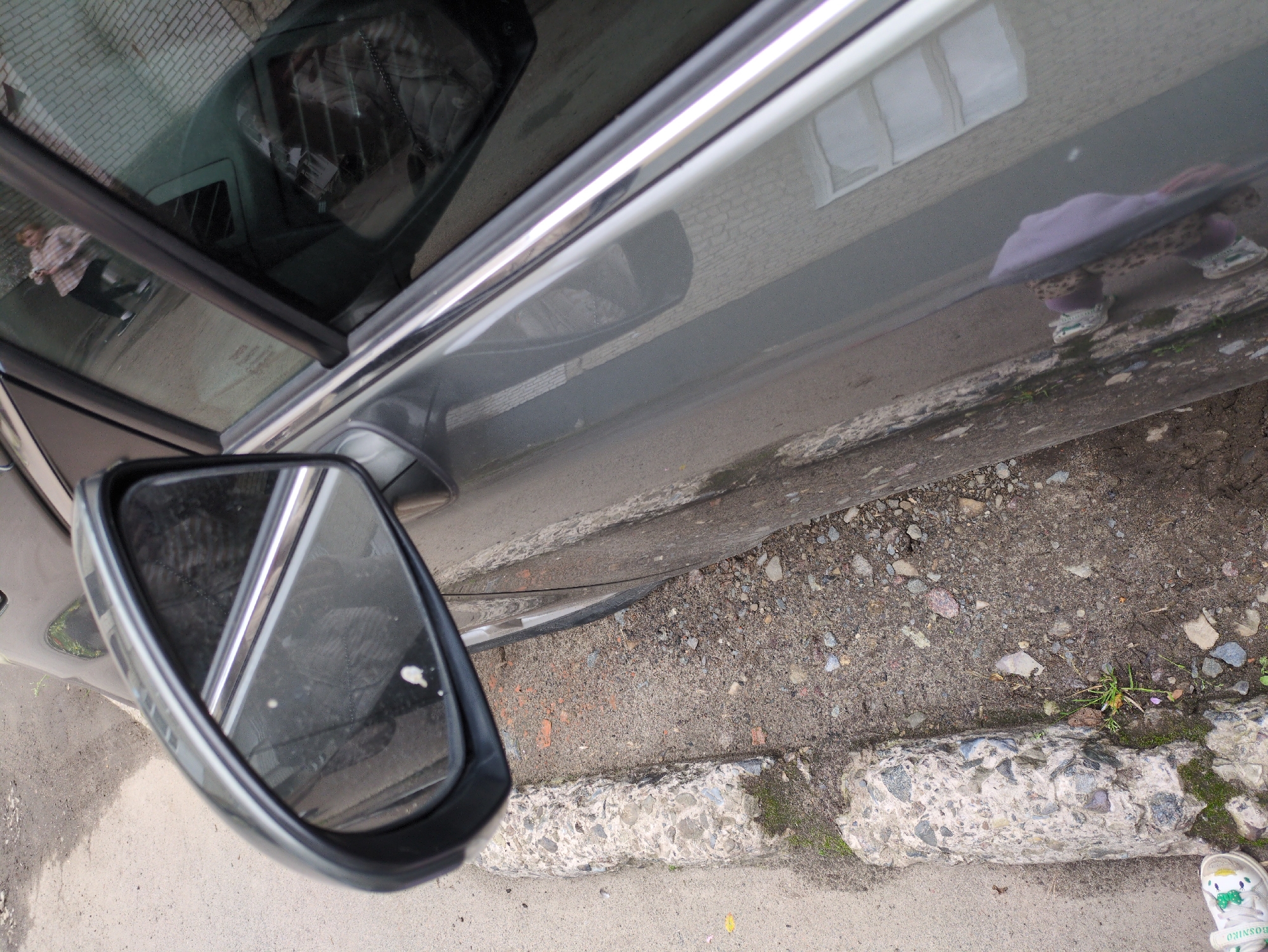 The image shows a close-up of a car parked on a paved surface, possibly a street or parking lot, next to a curb. The car is dark gray and reflects the surroundings. In the side mirror, a person in a plaid shirt and dark pants is visible, as well as a brick building. The ground is covered with dirt, small stones, and some sparse vegetation. A concrete curb runs along the edge of the paved area. The bottom left of the image shows a white sneaker with green accents. Based on the information provided, this scene is located in Dubliany, Ukraine.