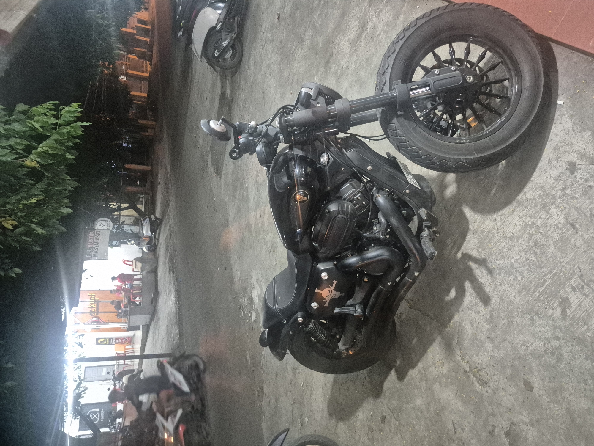 A black motorcycle with a skull and crossbones decal sits parked on a concrete surface. The motorcycle is the main subject. Other motorcycles are visible in the background, along with a building that has tables and chairs indicating a restaurant or cafe. People are seated at the tables. The scene is in Lhokseumawe, Indonesia, at night.