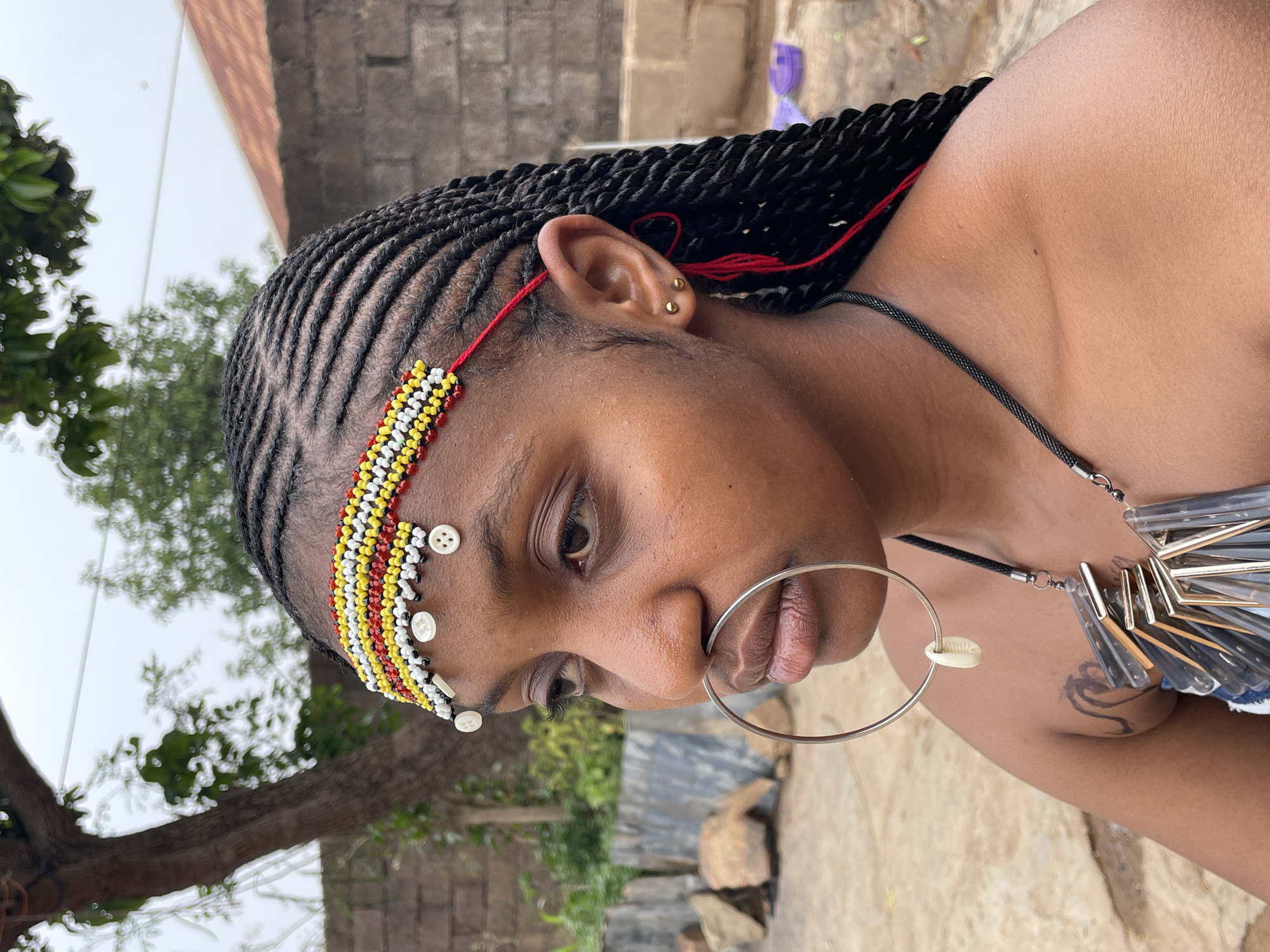 A close-up shot shows a woman with braided hair, adorned with a beaded headband featuring white buttons. She wears a large silver ring in her lip. A black cord necklace with a modern pendant of clear, silver, and blue materials and a single small shell is visible. The background suggests an outdoor setting, possibly in Jos, Nigeria, given the presence of a brick wall, tree branches, and a corrugated metal structure. The woman is looking downwards, and her expression is contemplative.