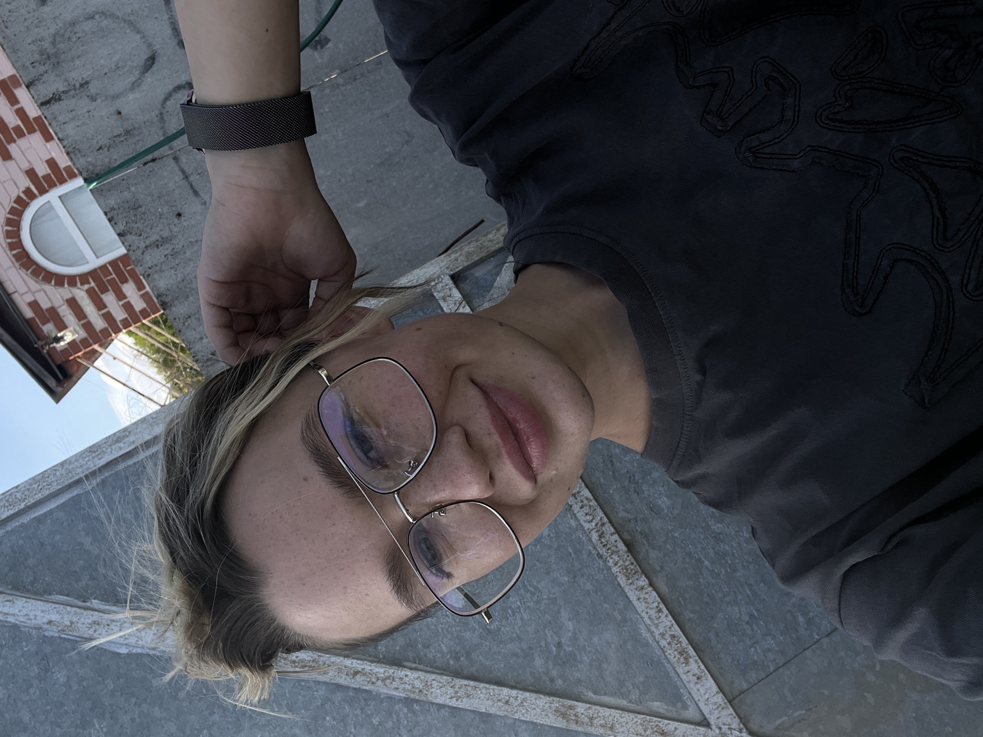 A young person with blonde hair and glasses is looking directly into the camera. The person is wearing a dark grey shirt with a decorative pattern. A metal watchband is visible on the person's wrist. In the background, there is a brick building with a curved window, suggesting an urban setting. The setting is possibly within the city of Слобода, Russia, based on the location information.