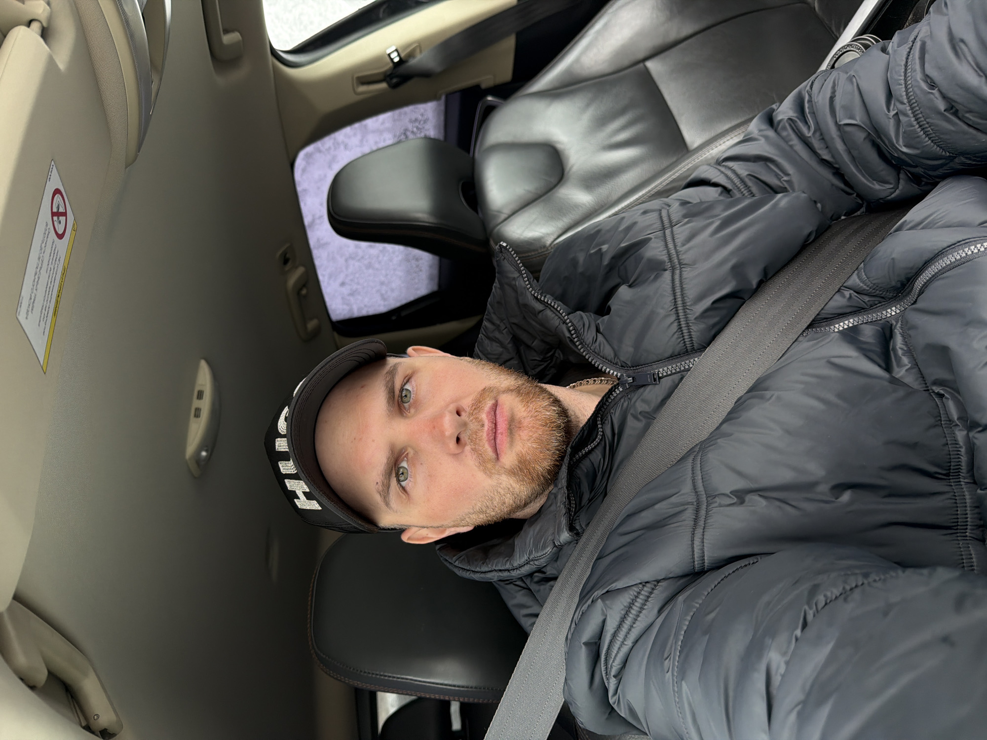 A man with a beard and wearing a black puffy jacket and a baseball cap is in the driver's seat of a car. He is wearing a seatbelt. The car is parked, and the scene appears to be from inside the vehicle. The location in the city of Chaikovsky, Russia is not identifiable from the image.