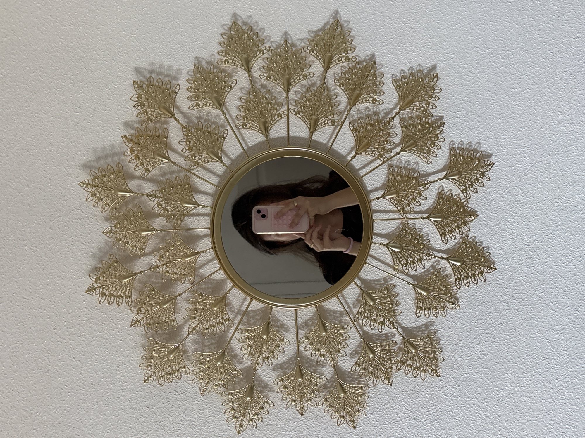 The image shows a golden decorative mirror mounted on a white, textured wall. The mirror has a sunburst design made of ornate, feather-like metal pieces. In the reflection of the mirror, a person is visible, holding a pink smartphone. The person's face is not fully visible, but their dark hair and hands are seen. The background of the reflection appears to be a room, with the white wall and mirror frame also reflected. The lighting suggests an indoor setting, possibly during the daytime given the brightness on the wall. There is no visible text in the image. The overall impression is a decorative wall accent in a domestic setting.