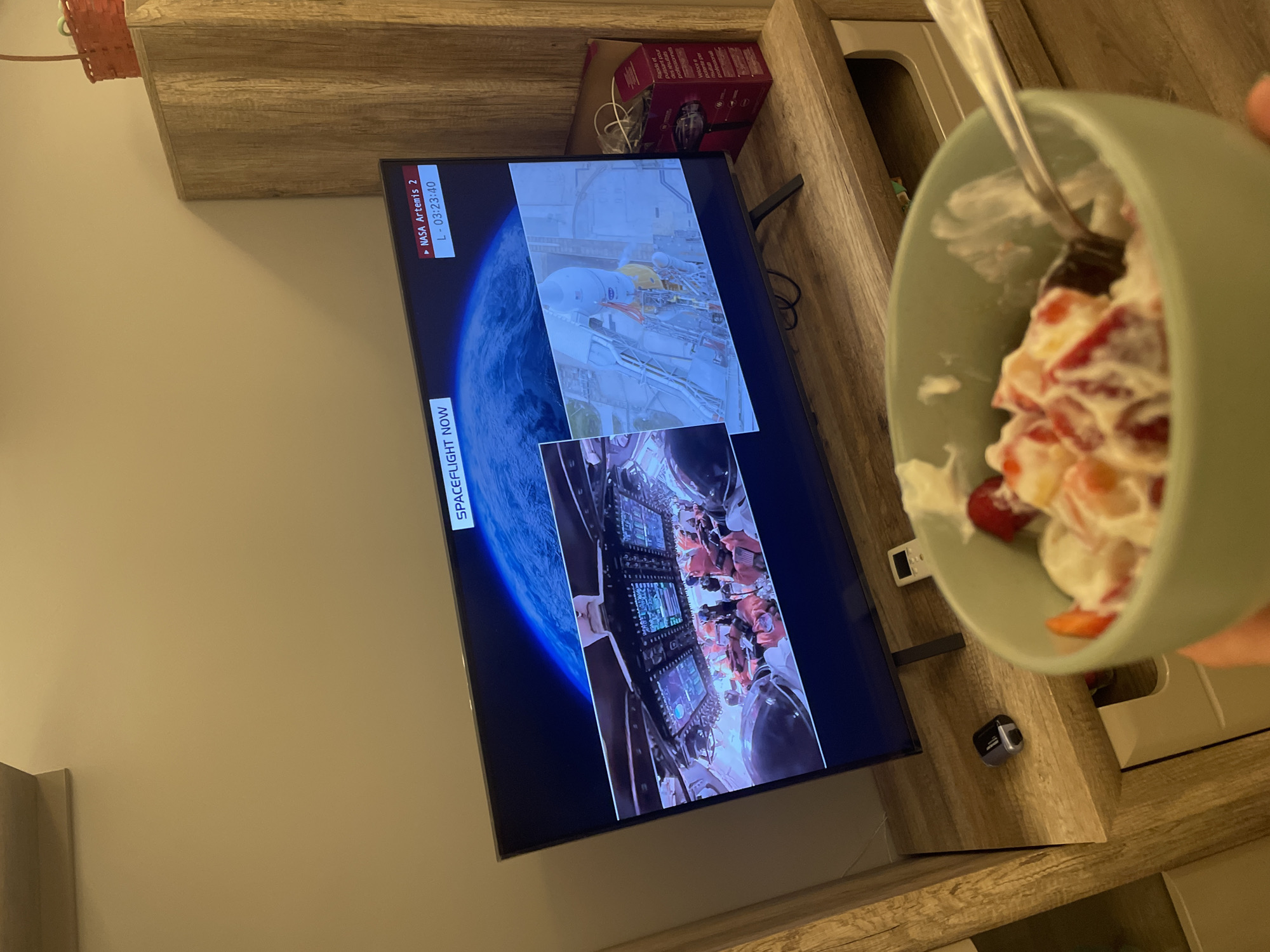 The image shows a television displaying a NASA Artemis 2 spaceflight broadcast, with a timer showing "L - 03:23:40." A bowl of fruit and cream is held in the foreground. The TV is situated on a light wood-grain cabinet. The background includes an adjacent wall painted in a neutral tone and a decorative woven basket on the top of the cabinet. There is no information to indicate the city of Sofia, Bulgaria.