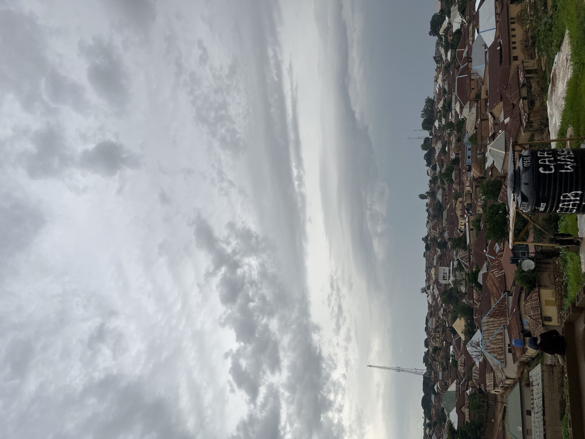This image depicts a densely populated hillside settlement in Jos, Nigeria. The scene is dominated by a multitude of buildings with predominantly corrugated iron roofs, showcasing a variety of colors from rust to silver and blue. The settlement sprawls across a steep incline, with structures seemingly built atop one another. Lush green vegetation is interspersed with the buildings, suggesting a natural environment that is being encroached upon by the urban development.
The sky is overcast with heavy, grey clouds, indicating a gloomy or potentially rainy weather condition. The time of day is unclear due to the lack of direct sunlight, but the overall lighting suggests it could be late afternoon or a heavily clouded day. A tall telecommunications tower is visible in the distance, adding a modern element to the otherwise traditional-looking settlement. In the foreground, on the right side of the image, a large black water tank is present. White text is painted on the tank, with "CAR WASH" and "OPEN" being partially legible. A person, mostly obscured, is visible near the water tank. The composition and the view from an elevated position suggest the photo was taken from a higher vantage point, overlooking the entire community.
