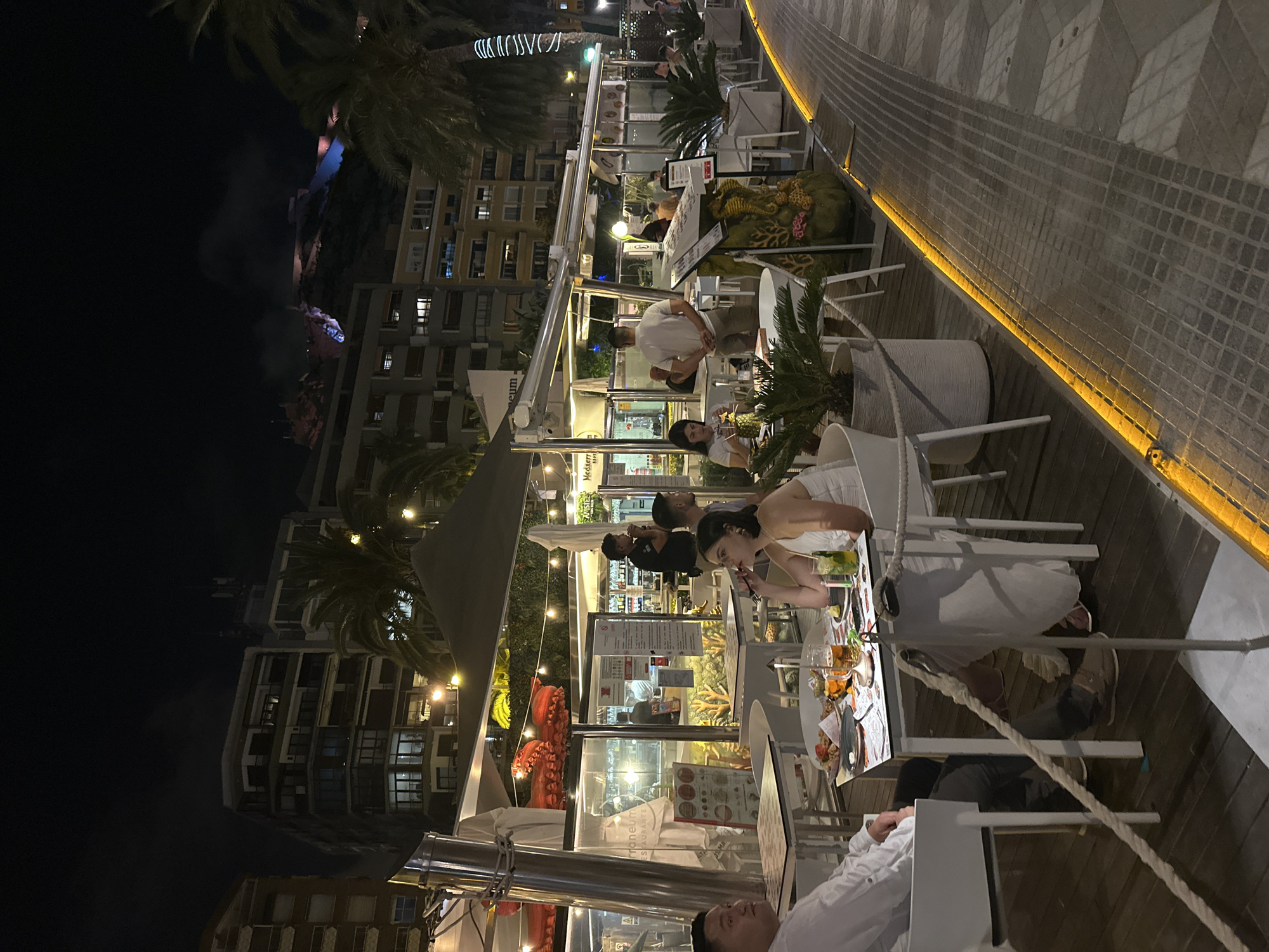 The image shows an outdoor seating area of a restaurant at night, with people dining at tables. The restaurant appears to be located in a city with multi-story buildings in the background, and palm trees are visible. The tables are set with food and drinks, and string lights illuminate the scene. A couple is seated at a table in the foreground, with a man on the left and a woman on the right. The man is wearing a white shirt and dark pants, and the woman is wearing a white dress. Other diners and restaurant staff are visible in the background. The ground is paved with tiles, and a rope barrier is present. The scene is lit by artificial lights, creating a warm and inviting atmosphere.