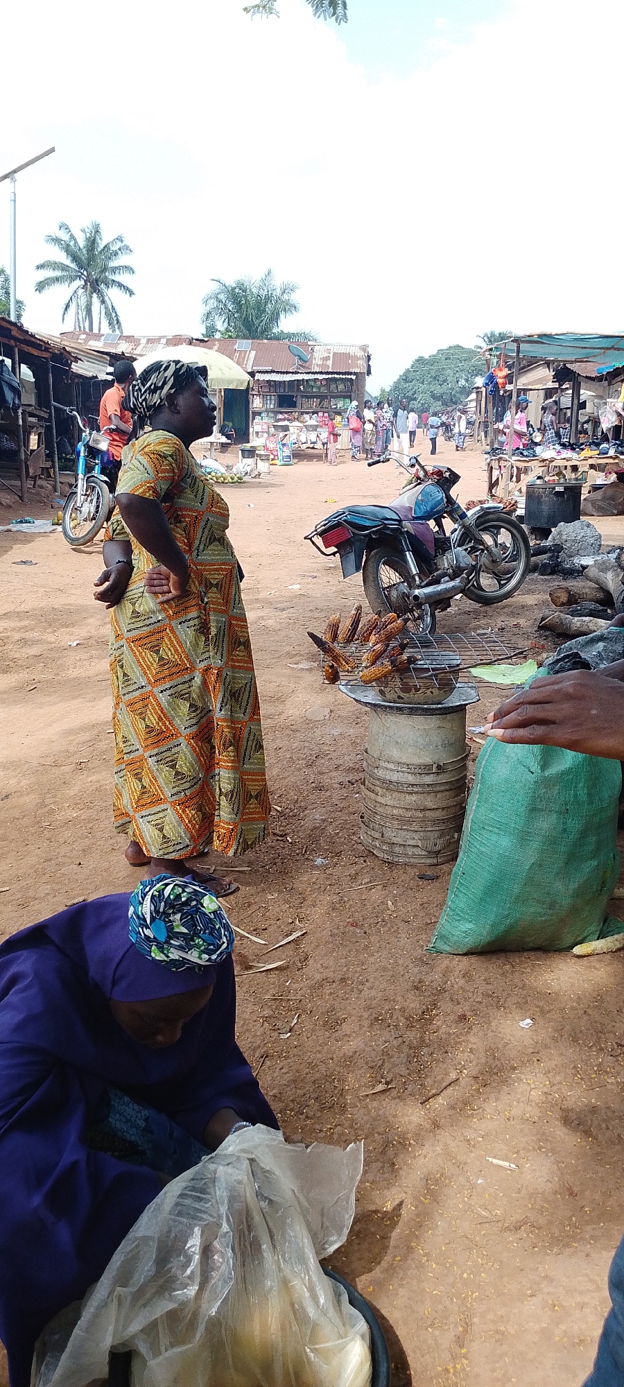 This is a full-shot, eye-level, vertical photograph taken outdoors, likely in a market setting in Gumel, Nigeria. The foreground is dominated by a woman in a purple headscarf and blue robe, crouching and handling a large, clear plastic bag containing pale yellow goods. To her right, a hand reaches out from the right side of the frame. Behind the crouching woman, a woman stands with her hands on her hips, wearing a brightly patterned, long dress and a headscarf. She is looking to her right.
In the mid-ground, there is a motorcycle parked on the right, and to its left, a grill with several ears of corn cooking over a fire. The grill is placed on top of a stack of what appear to be industrial metal barrels. The background shows a dusty, unpaved market street lined with stalls and small buildings with corrugated metal roofs. Several other people are visible in the background, some appearing to be customers and vendors. A solar-powered street lamp is visible on the far left. The sky is overcast with a bright sky.