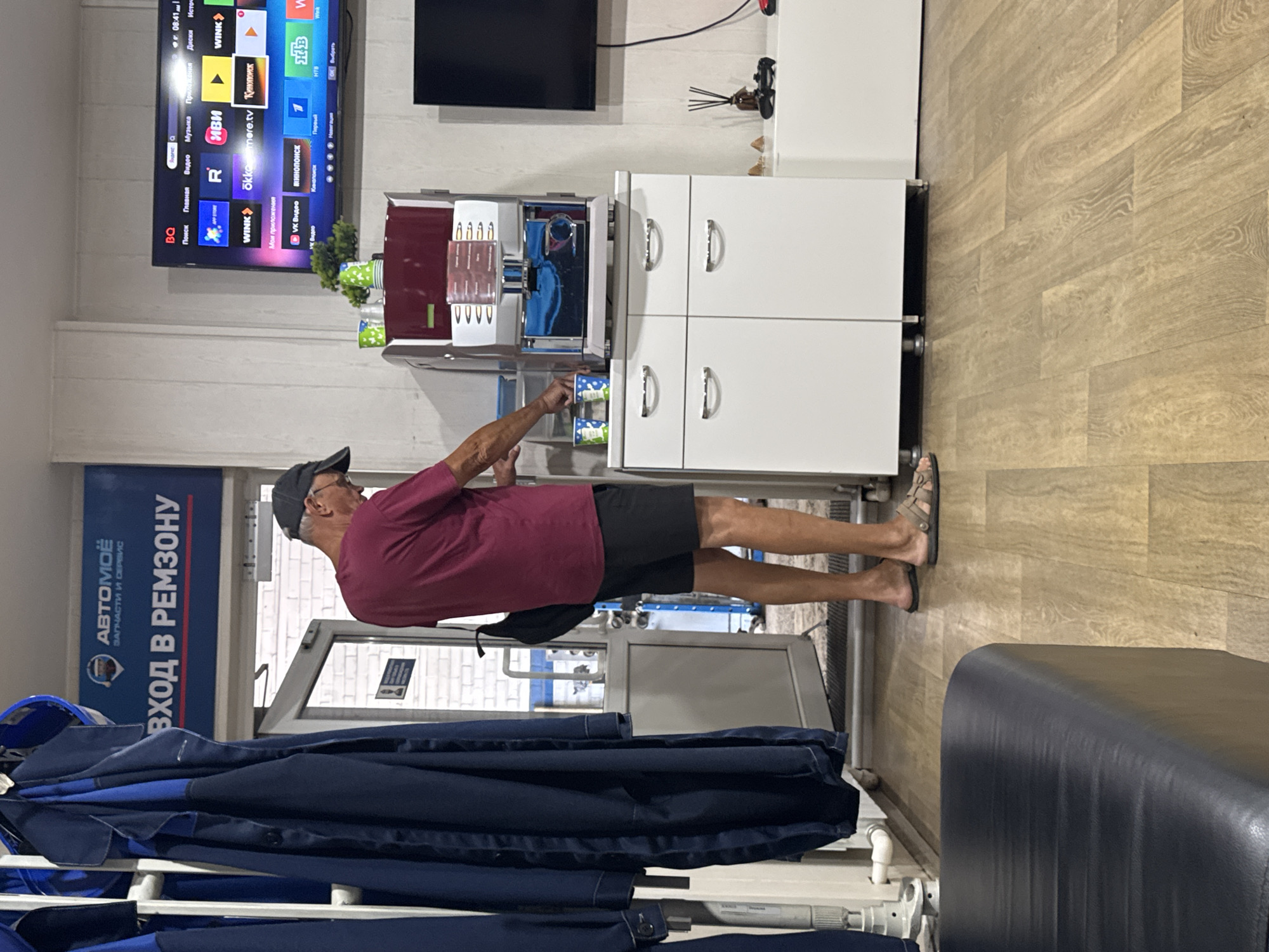 A man in a burgundy t-shirt and black shorts stands in front of a white cabinet with a coffee machine on top. He is reaching for the coffee machine, and there are two cups on the counter next to it. The background shows a doorway with a sign that reads "ВХОД В РЕМЗОНУ" (Entrance to the repair zone) with the logo of "АВТОМОЁ" (Avtomoe). A TV screen is visible on the right side of the image, displaying a variety of icons for streaming services. The flooring appears to be light-colored wood laminate. The scene is indoors, possibly in a service center or waiting area. This location is in the city of Chaikovsky, Russia.