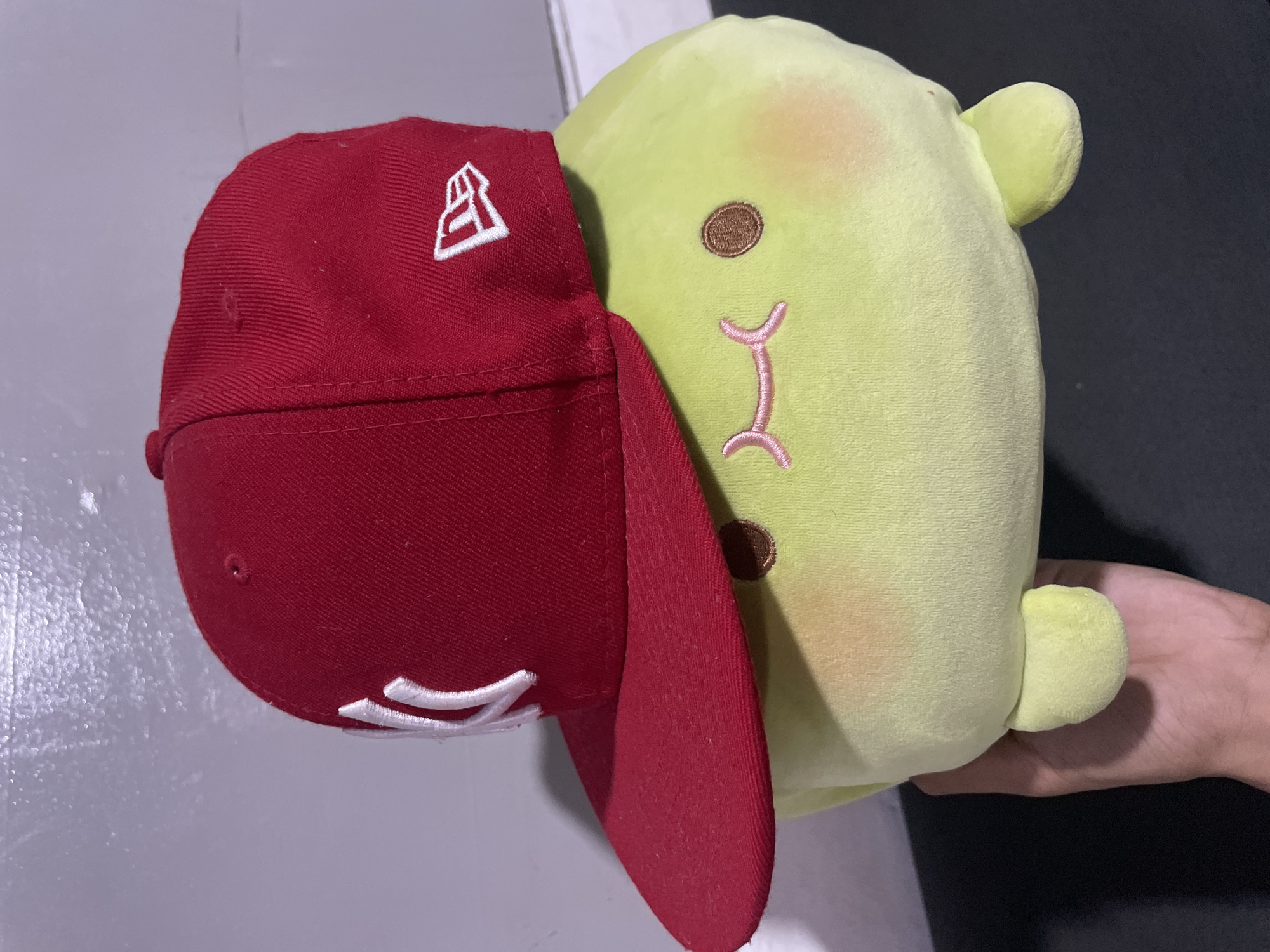 An indoor scene features a light green, plump plush toy, resembling a blob or a large pea, being held by a human hand. The plush toy has two small, dark brown embroidered eyes and a pink, inverted 'm'-shaped embroidered mouth, giving it a gentle expression, along with subtle orange blush on its 'cheeks' and two small, rounded foot-like appendages at its base. It is wearing a red New York Yankees flat-brimmed baseball cap, with the white 'NY' logo prominently embroidered on the front and a white New Era flag logo on the wearer's right side. The cap shows some minor lint or dust. The human hand, with light to medium brown skin, supports the toy from beneath. The background consists of a light grey wall with a slightly mottled or reflective texture, and a dark, matte surface is visible beneath the hand and toy.