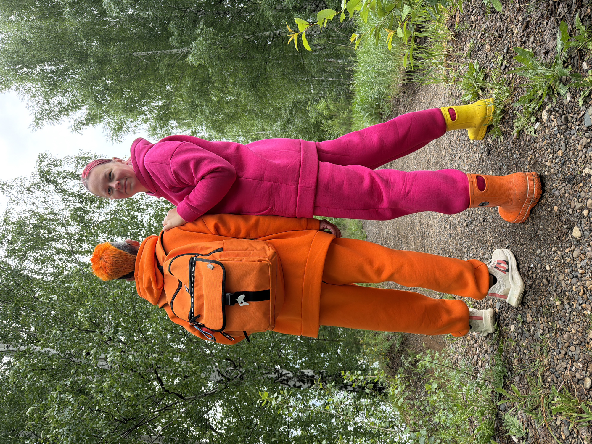 Two people in brightly colored clothing are positioned on a gravel path, likely in a park or natural area based on the presence of trees and vegetation. The person closer to the camera is wearing an orange hooded jacket and pants, with an orange backpack. They are wearing sneakers with red accents. The second person, positioned behind the first, wears a pink hooded jacket and pants, along with yellow boots. The background consists of trees and foliage. The location is specified as Chaikovsky, Russia.